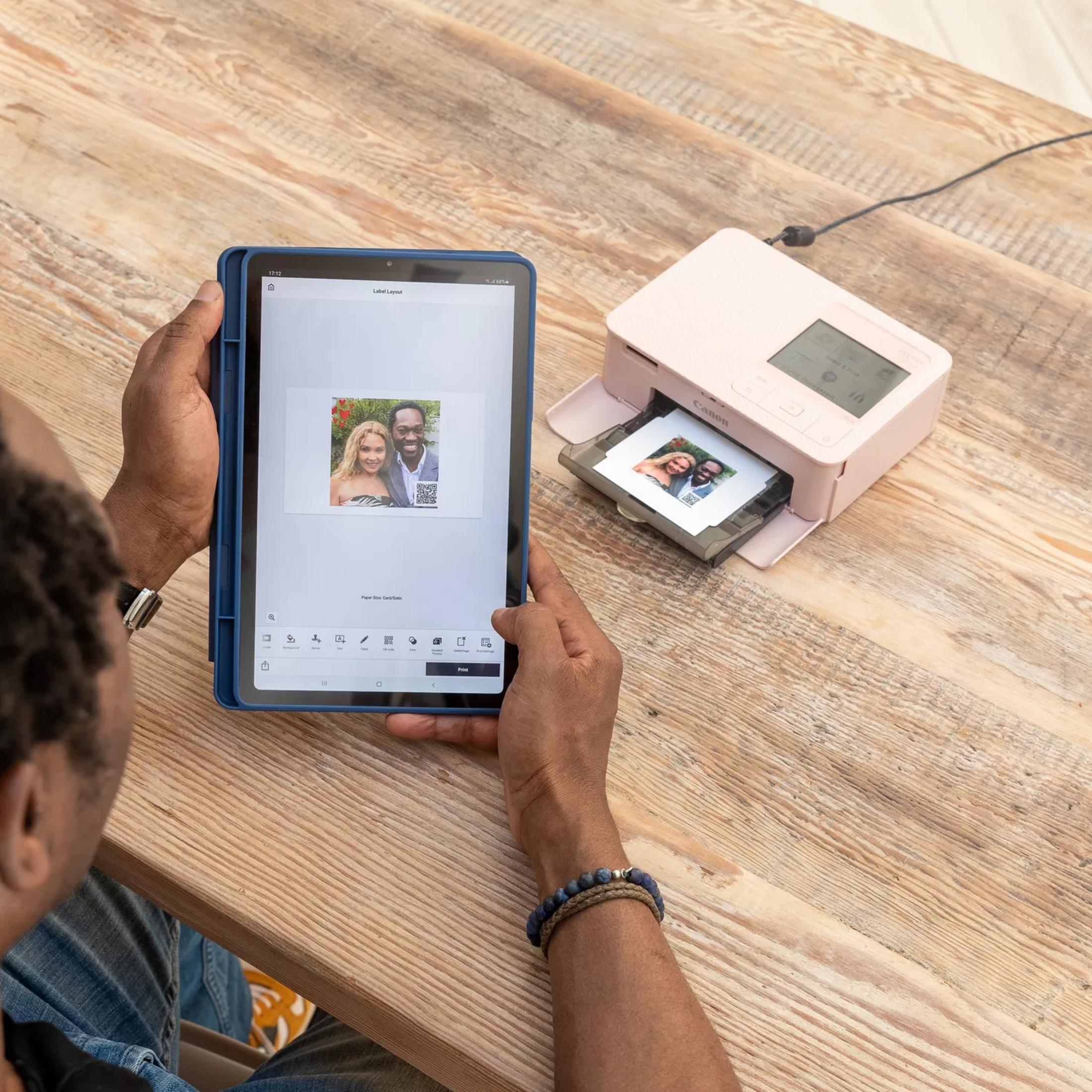Mężczyzna drukuje zdjęcie z tabletu na różowej drukarce przy drewnianym stole.