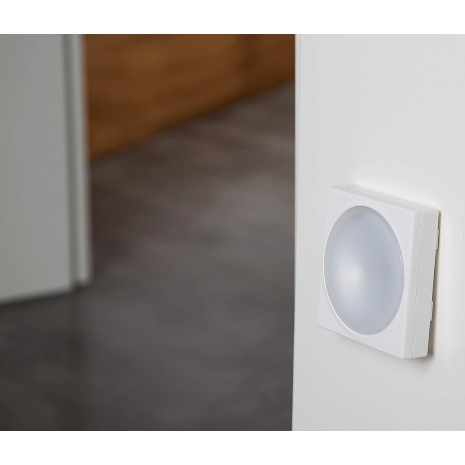 Ein quadratischer weißer Bewegungsmelder mit rundem, milchigem Lichtsensor an einer weißen Wand.