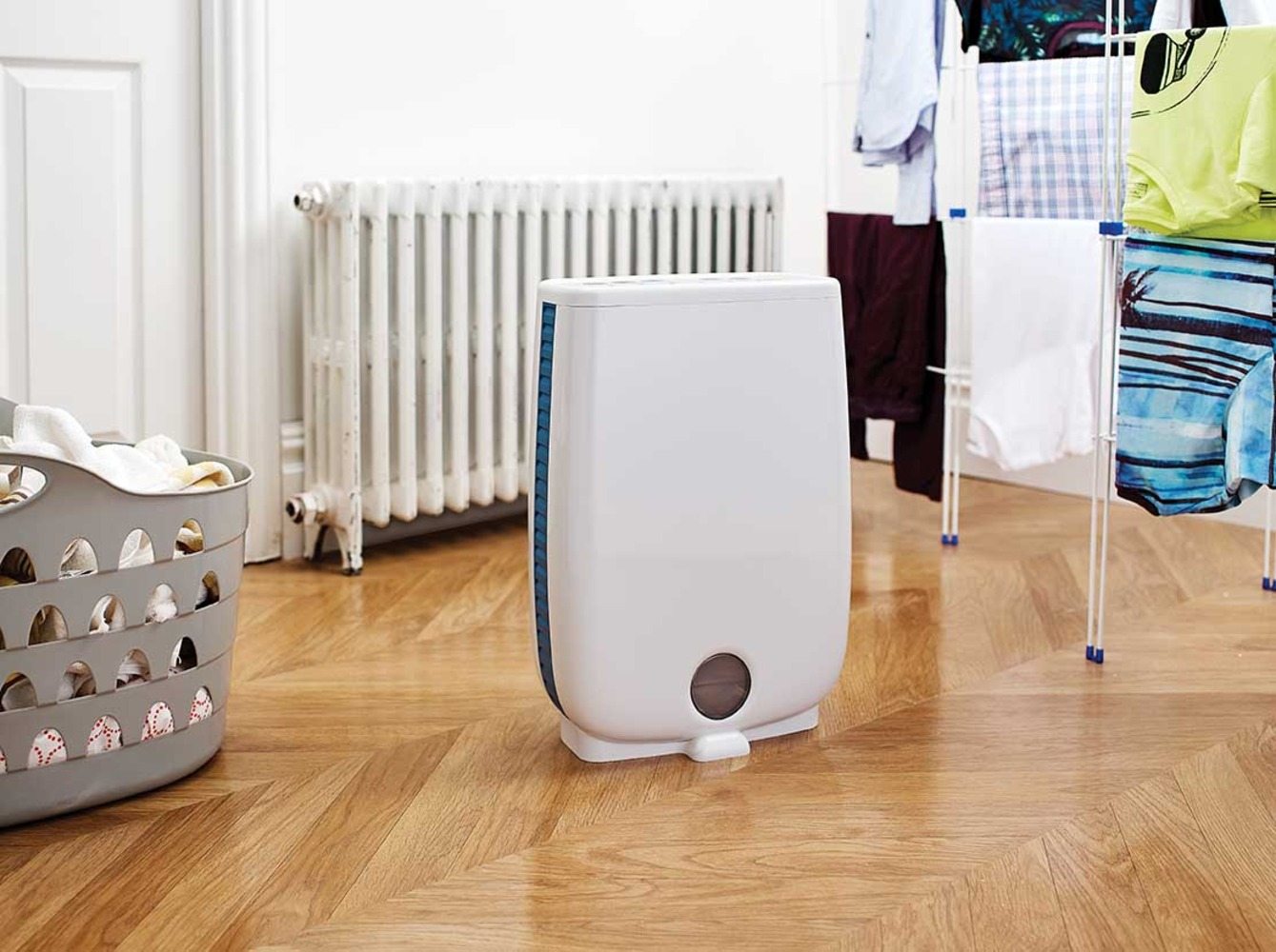 A white dehumidifier sits on a wooden floor next to a laundry basket and drying rack.