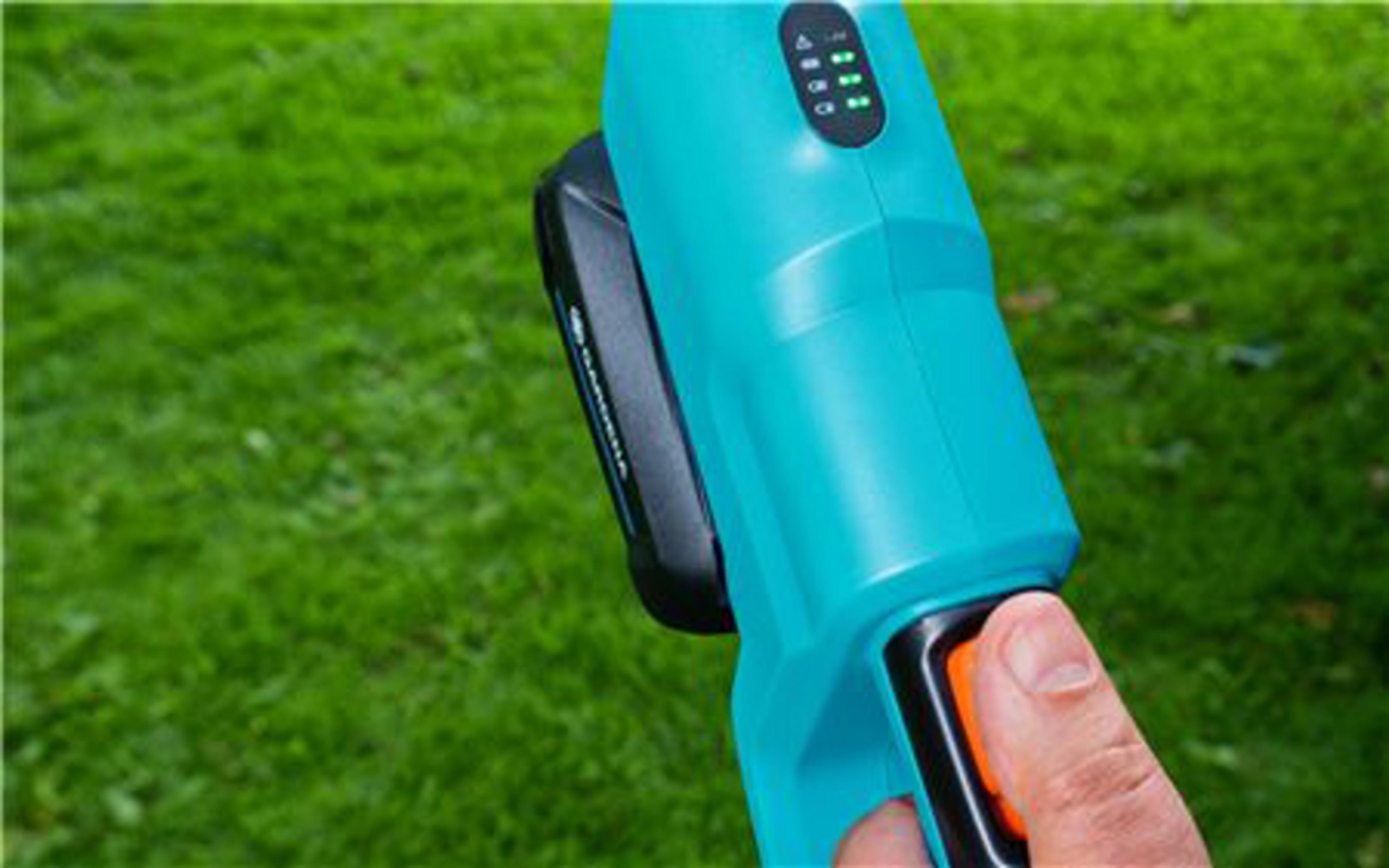 Eine Hand drückt einen orangefarbenen Knopf an einem tealfarbenen Gerät mit Batteriestandanzeige.