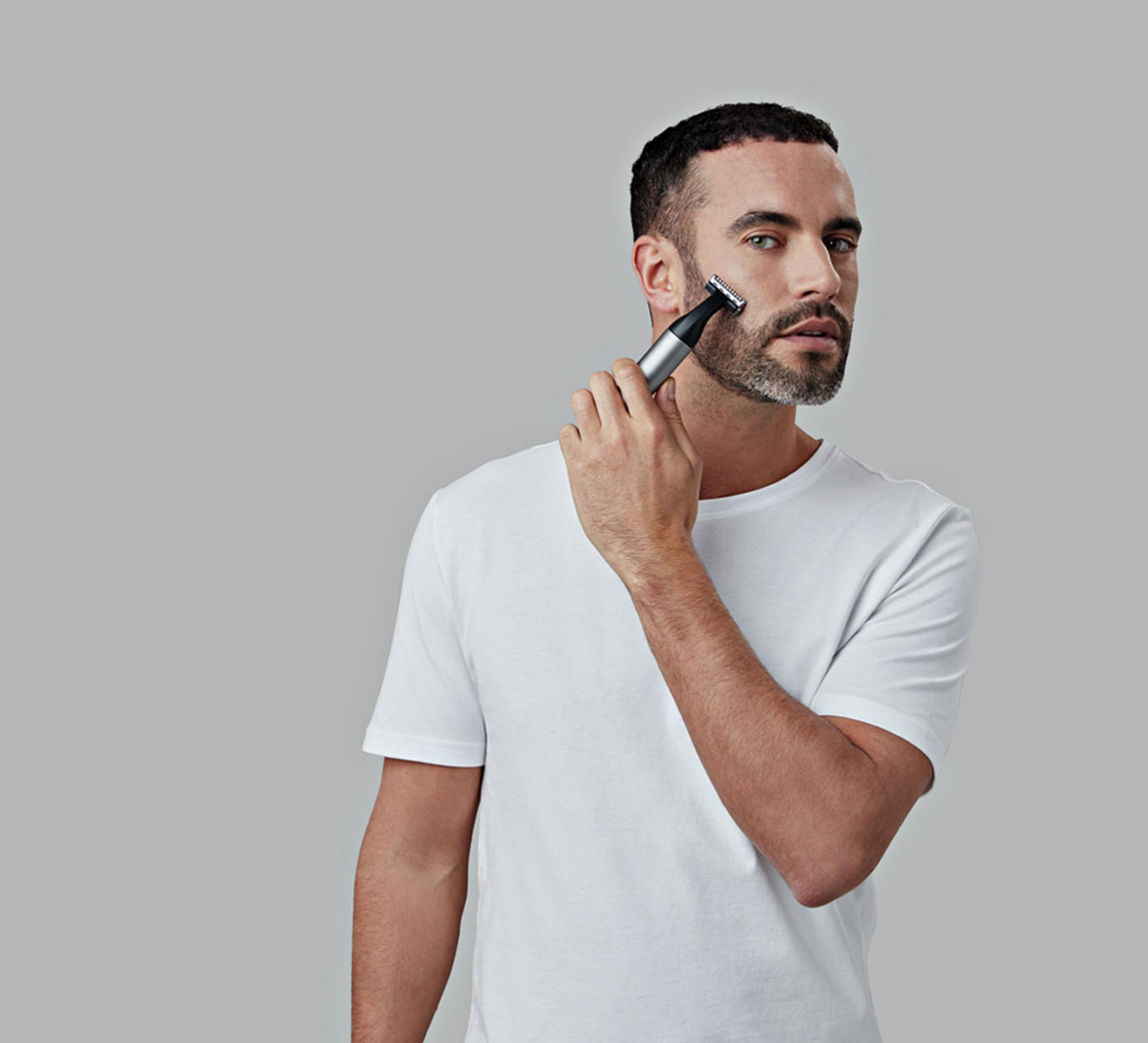 Mann mit Bart rasiert sich das Gesicht mit einem elektrischen Trimmer. Er trägt ein weißes T-Shirt.