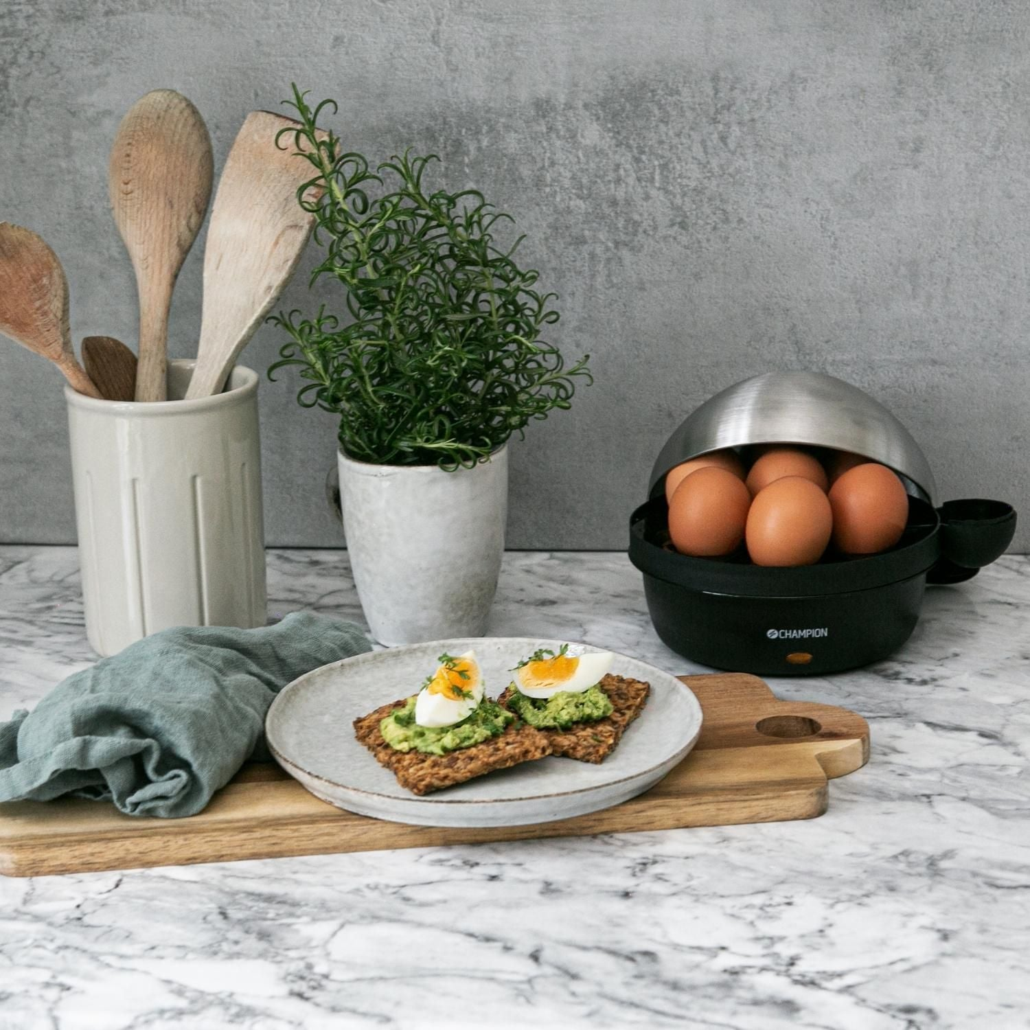 Egg cooker with eggs, herbs, and avocado toast on marble countertop.