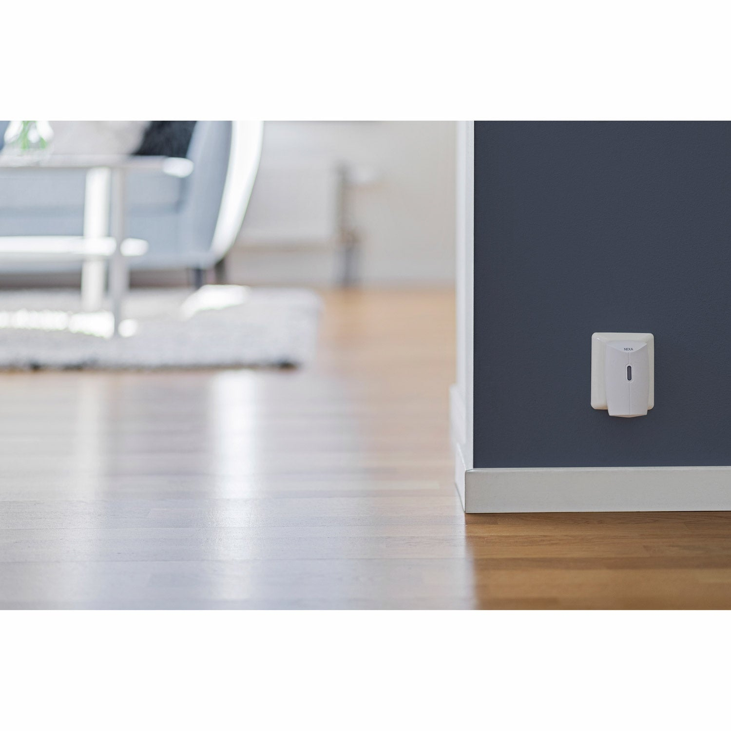 A white Nexa device on a dark blue wall above a wooden floor.