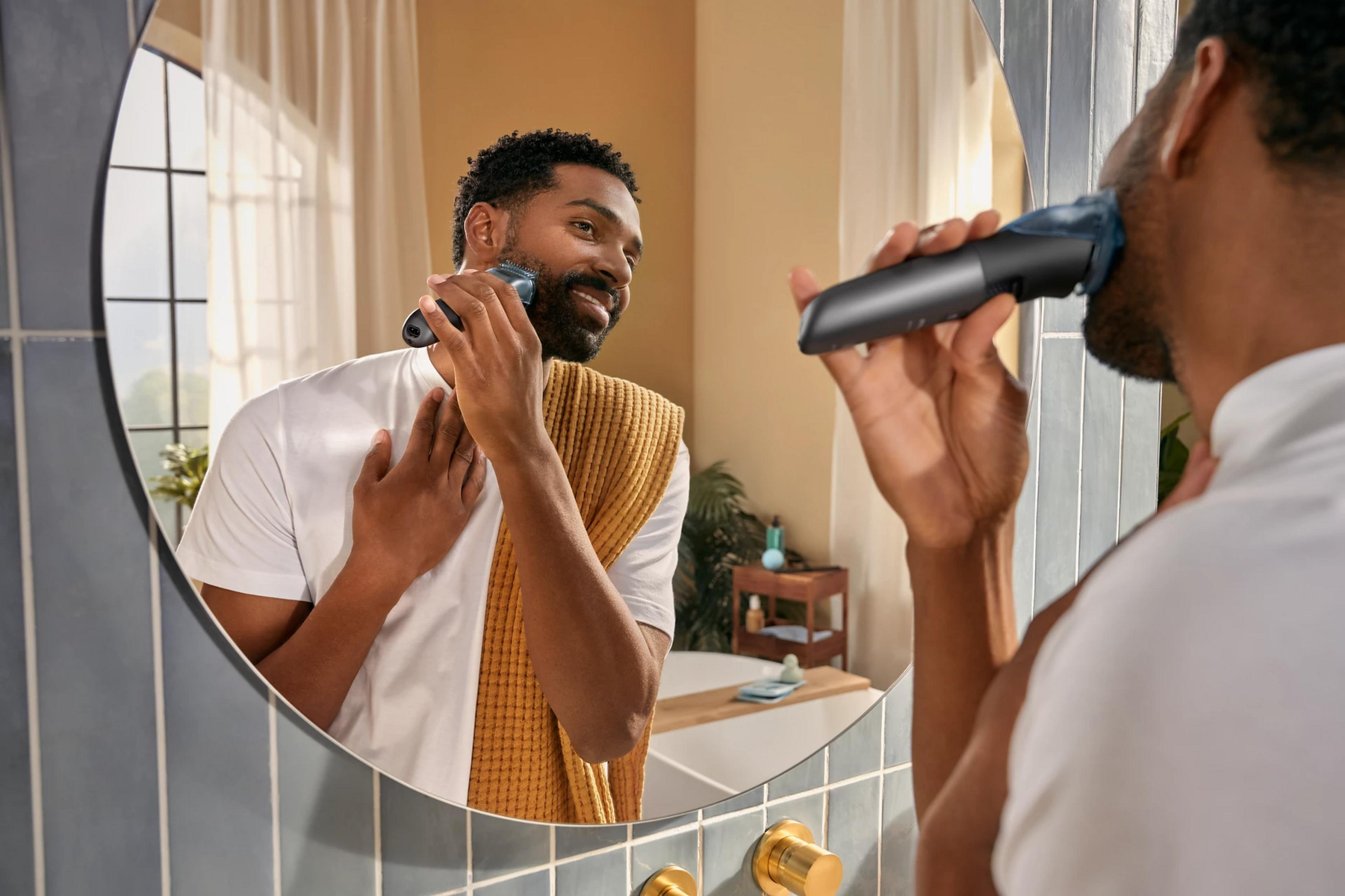 Mężczyzna goli twarz trymerem w lustrze. Nosi biały T-shirt.