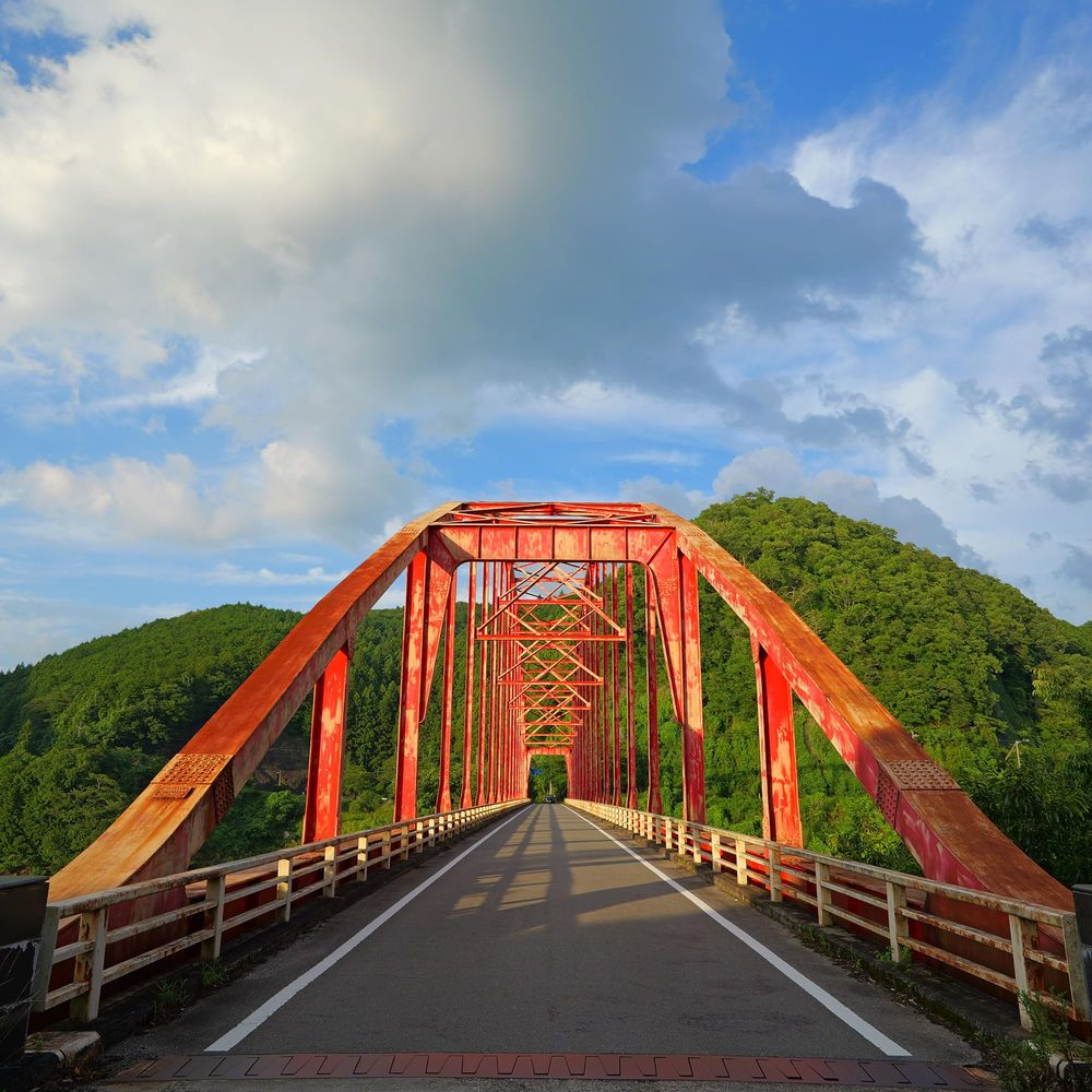 Un ponte ad arco rosso attraversa una strada verso una collina boscosa sotto un cielo blu.
