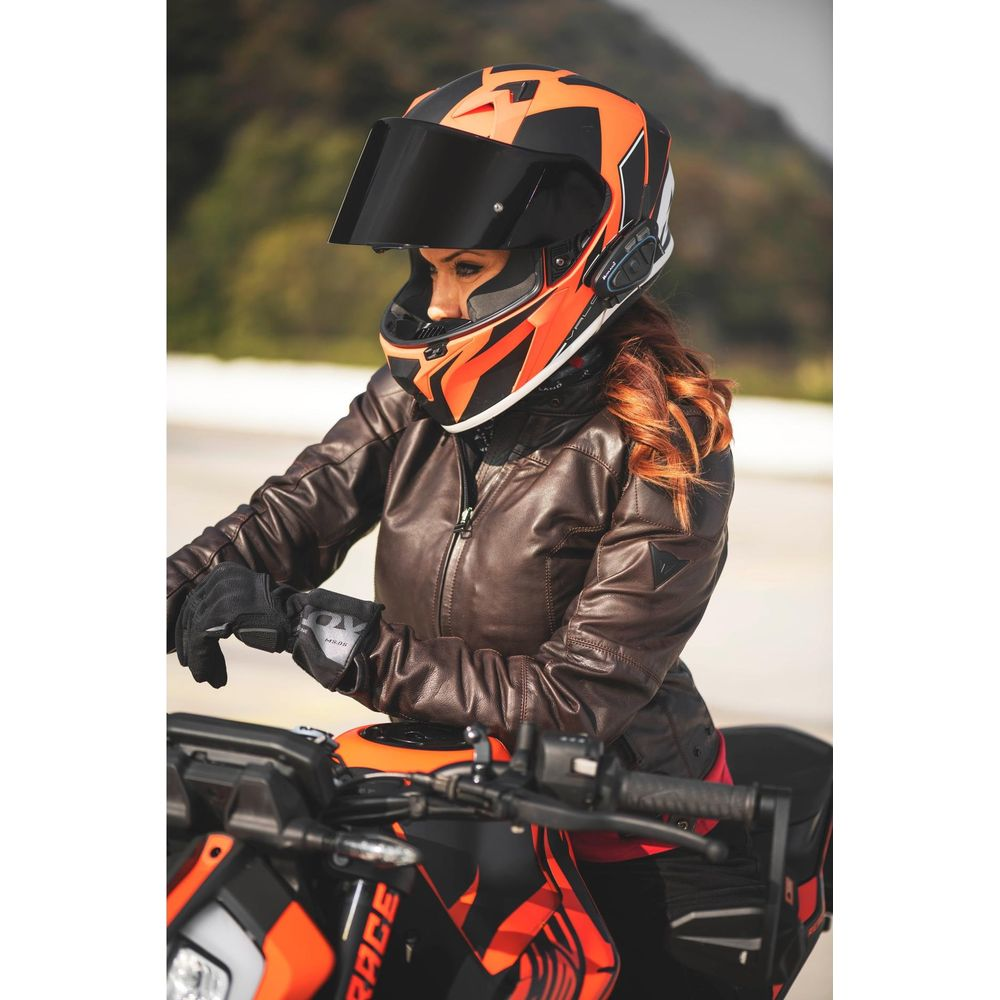 Kobieta w brązowej skórzanej kurtce i pomarańczowo-czarnym kasku siedzi na motocyklu.