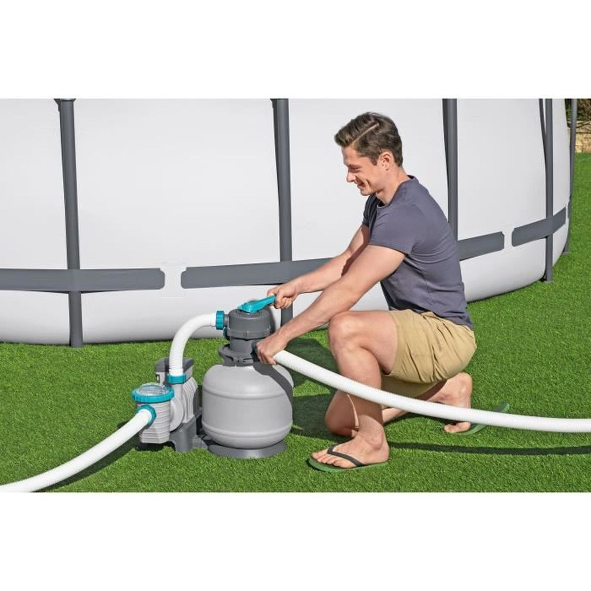 Mężczyzna klęczy przy filtrze basenowym, podłączając wąż. Zielona trawa.