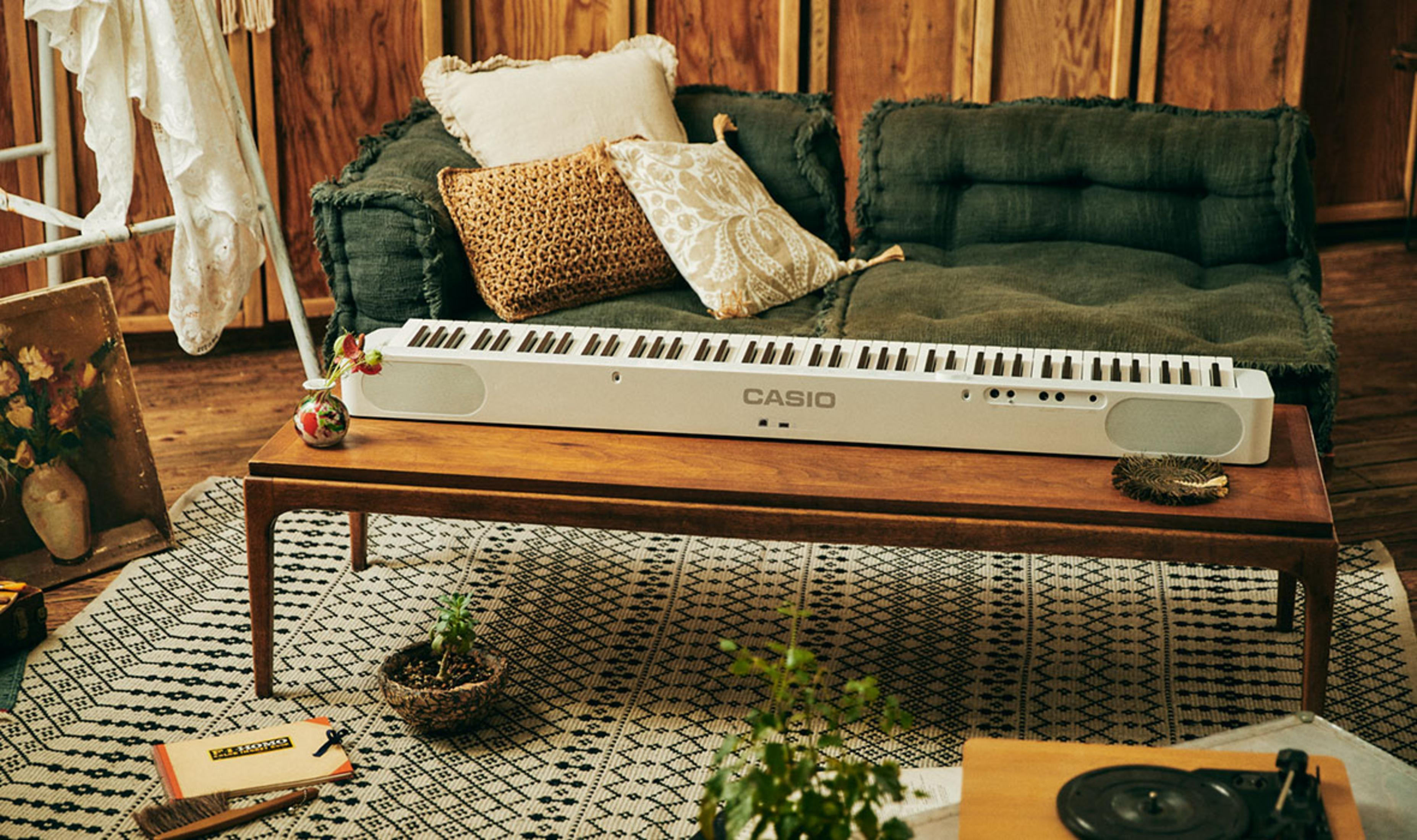 Ein weißes Casio Keyboard steht auf einem Holztisch, daneben ein Sofa mit Kissen.
