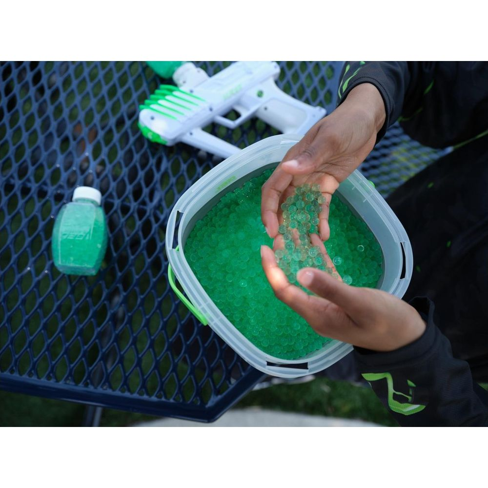 Manos sostienen bolitas verdes sobre un cubo; pistola de agua y botella cerca.