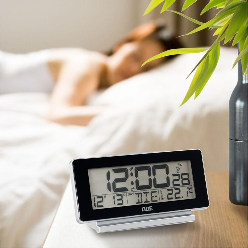 Digitaluhr zeigt 12:00 an, Person schläft im Hintergrund.