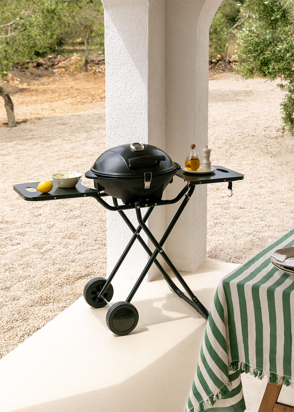 Una parrilla negra sobre un soporte con ruedas, junto a una mesa con mantel verde.