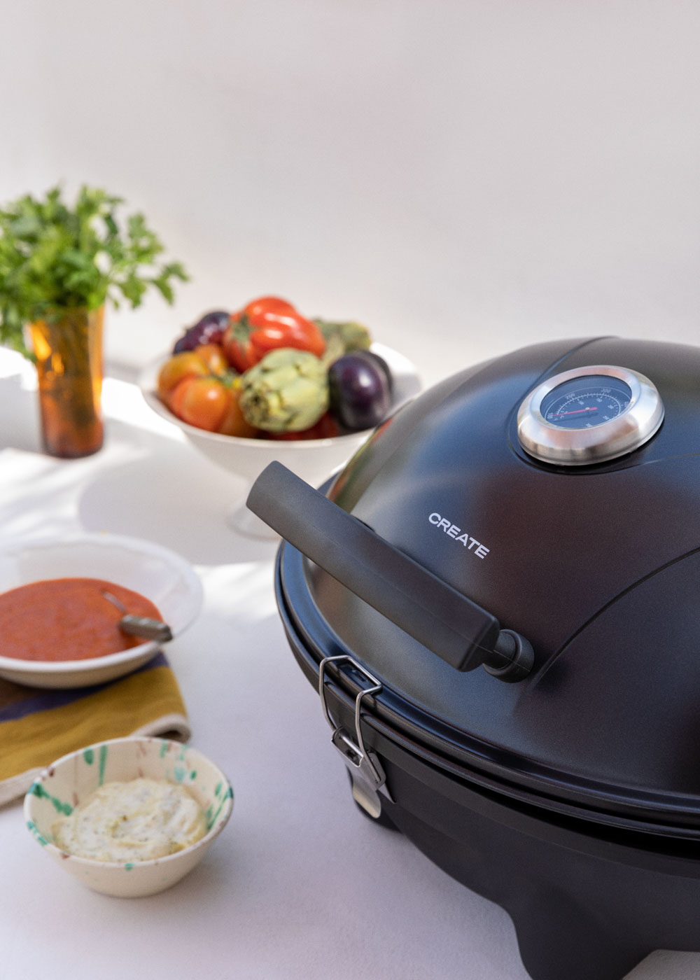 Una parrilla negra con termómetro, al lado de cuencos con verduras, salsa y dip.