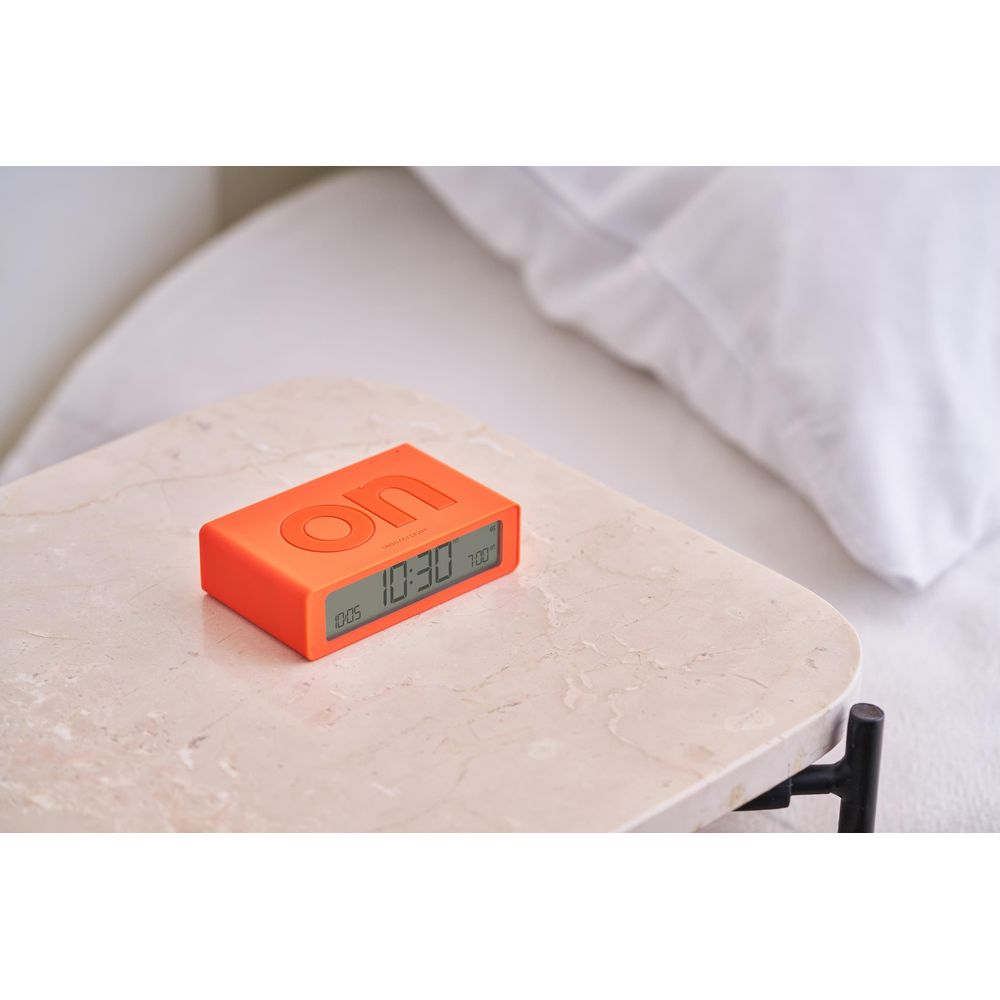 Un réveil numérique orange avec 'on' imprimé dessus est posé sur une table en marbre à côté d'un lit.