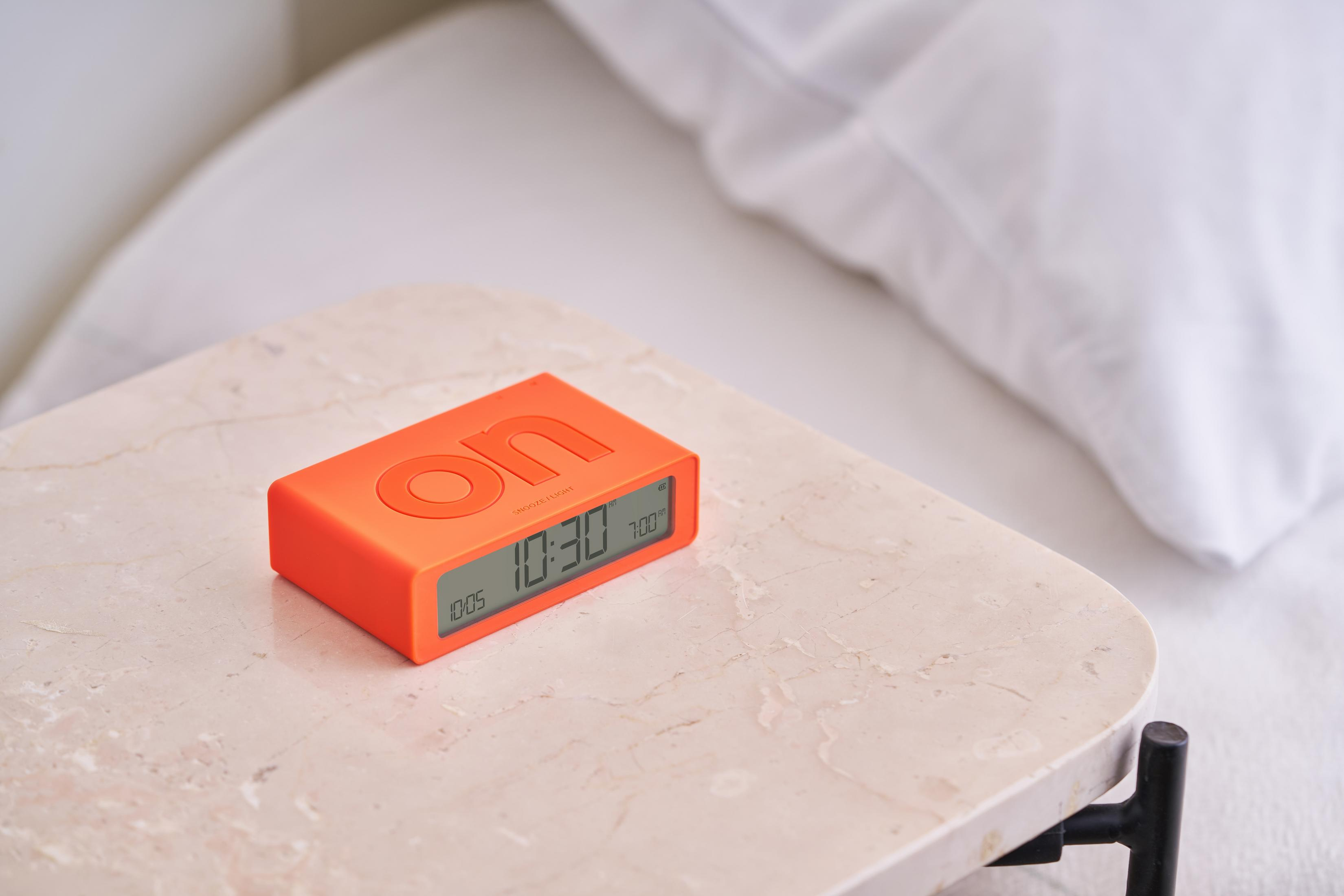 Un réveil numérique orange sur une table de chevet en marbre à côté d'un lit.