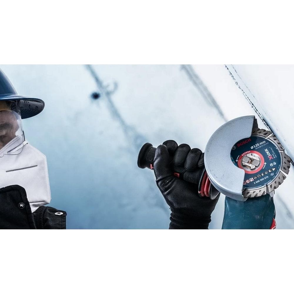 Mann mit Schutzbrille, Maske und Handschuhen hält Winkelschleifer an Wand.