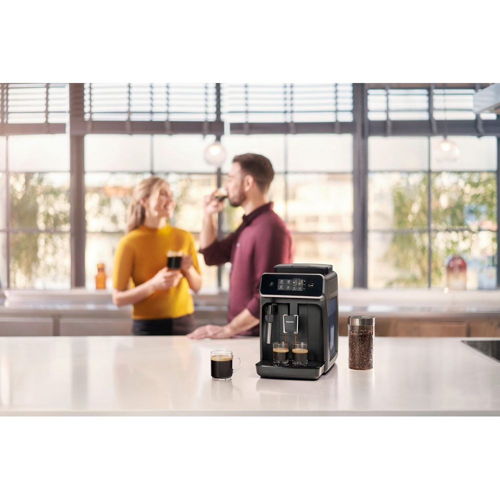 Kaffeemaschine mit zwei Espressotassen, Mann und Frau im Hintergrund trinkt.