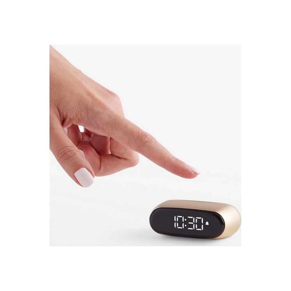 Eine Hand mit hellrosa Nägeln zeigt auf einen kleinen digitalen Wecker mit der Anzeige 10:30.