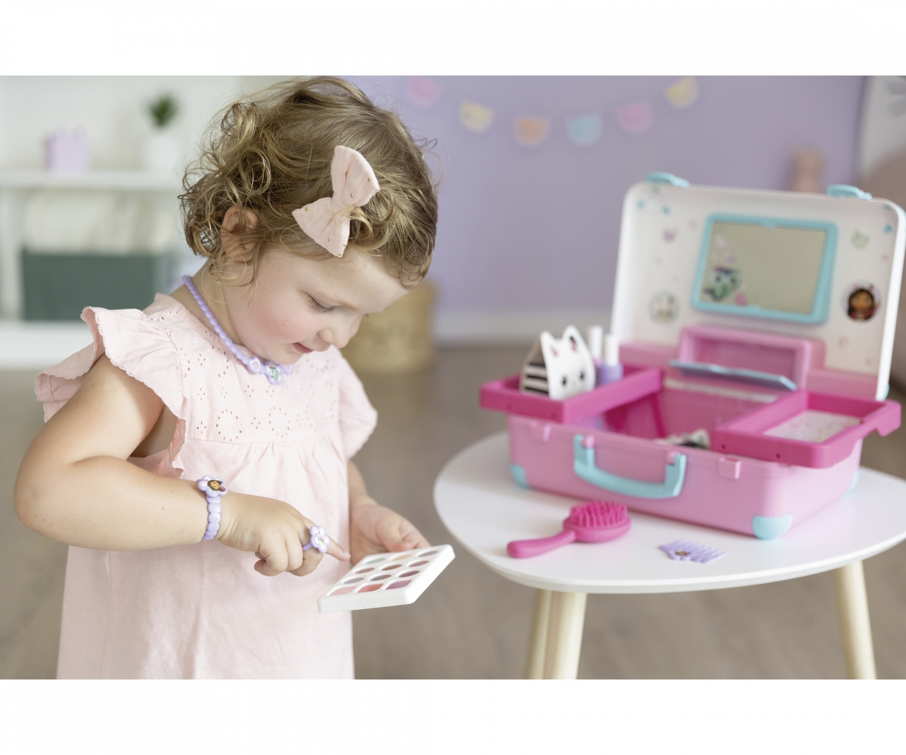 Une petite fille en robe rose joue avec une trousse de maquillage et des accessoires.
