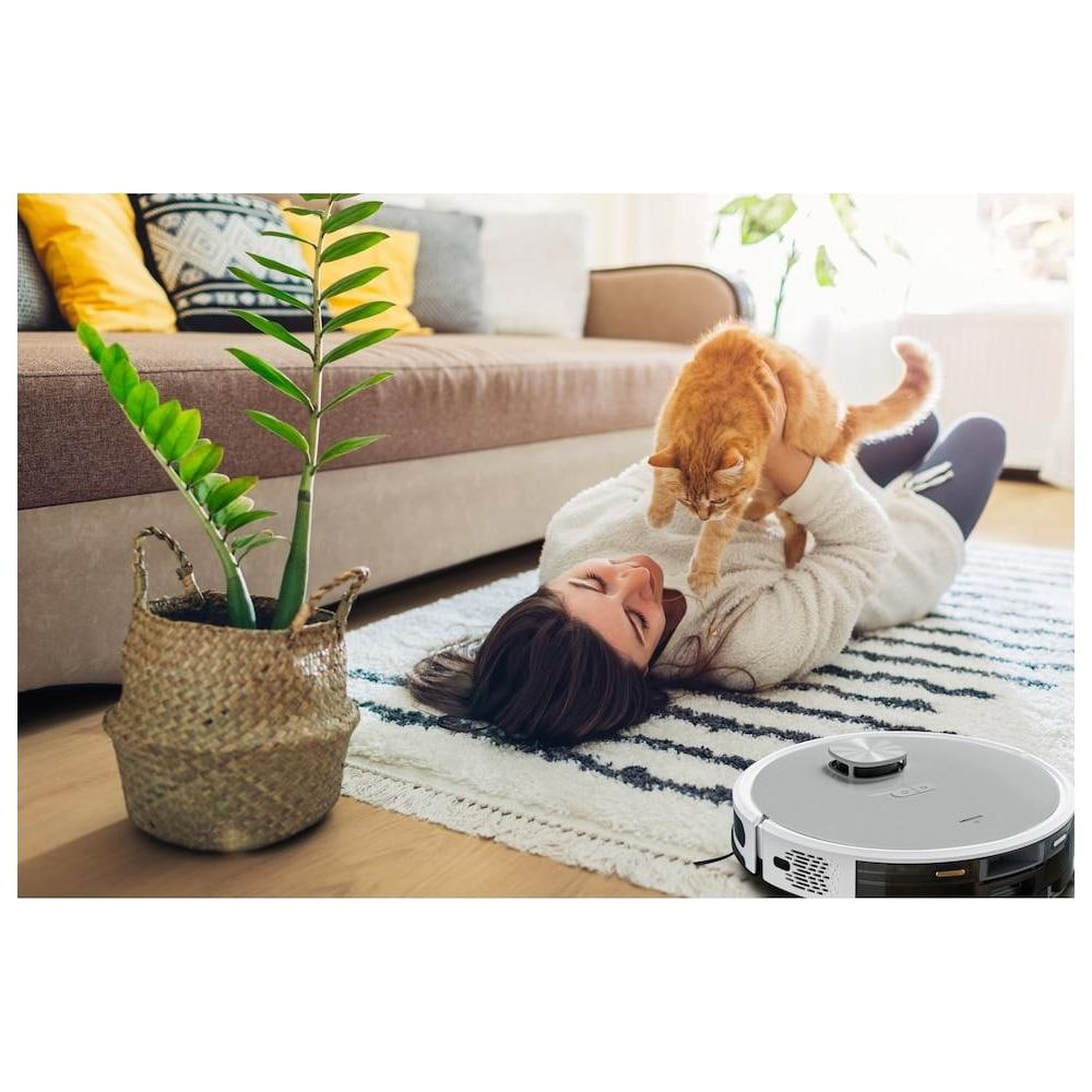 Kobieta leży na dywanie z kotem. Po prawej stronie znajduje się robot sprzątający.