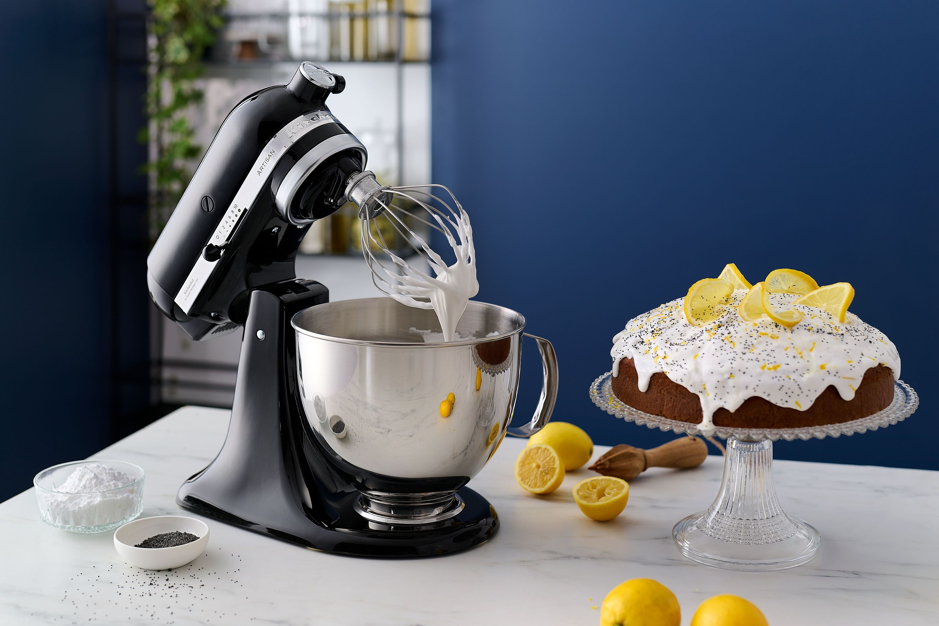 Ein schwarzer Küchenmixer verquirlt weiße Creme in einer Edelstahlschüssel neben einem Kuchen.