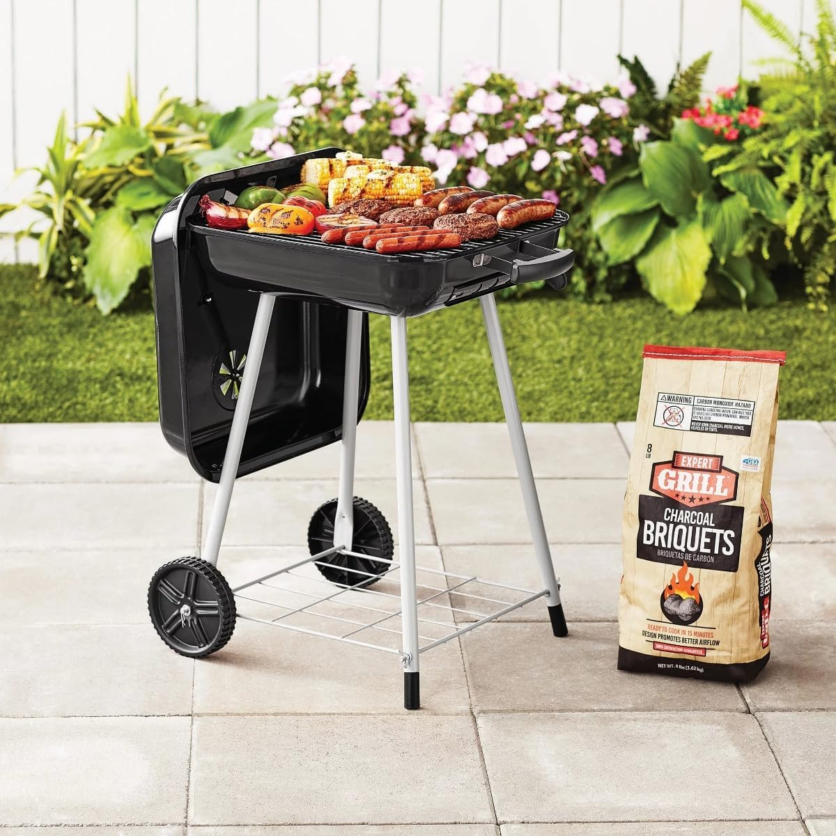 Ein Grill mit Würstchen, Mais und Gemüse steht auf einer Terrasse neben einer Kohlepackung.