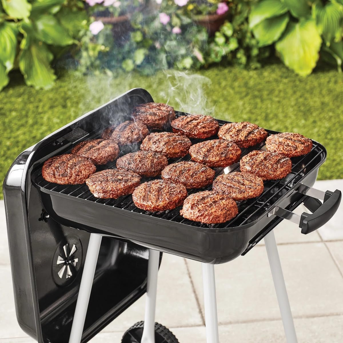 Viele Burger-Patties garen auf einem Holzkohlegrill in einem Garten.