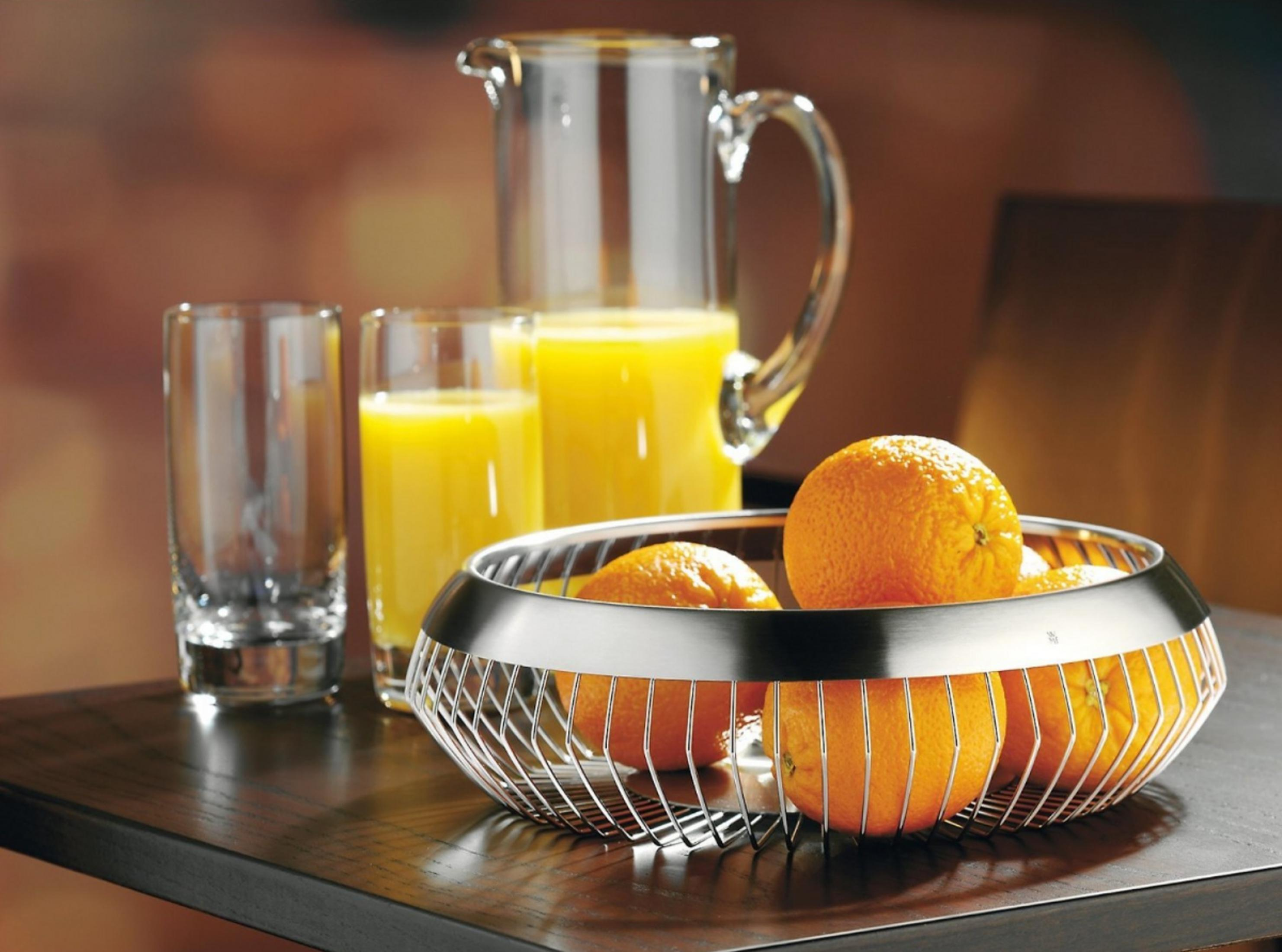 Orangen in einem Drahtkorb neben Gläsern und einem Krug mit Orangensaft.