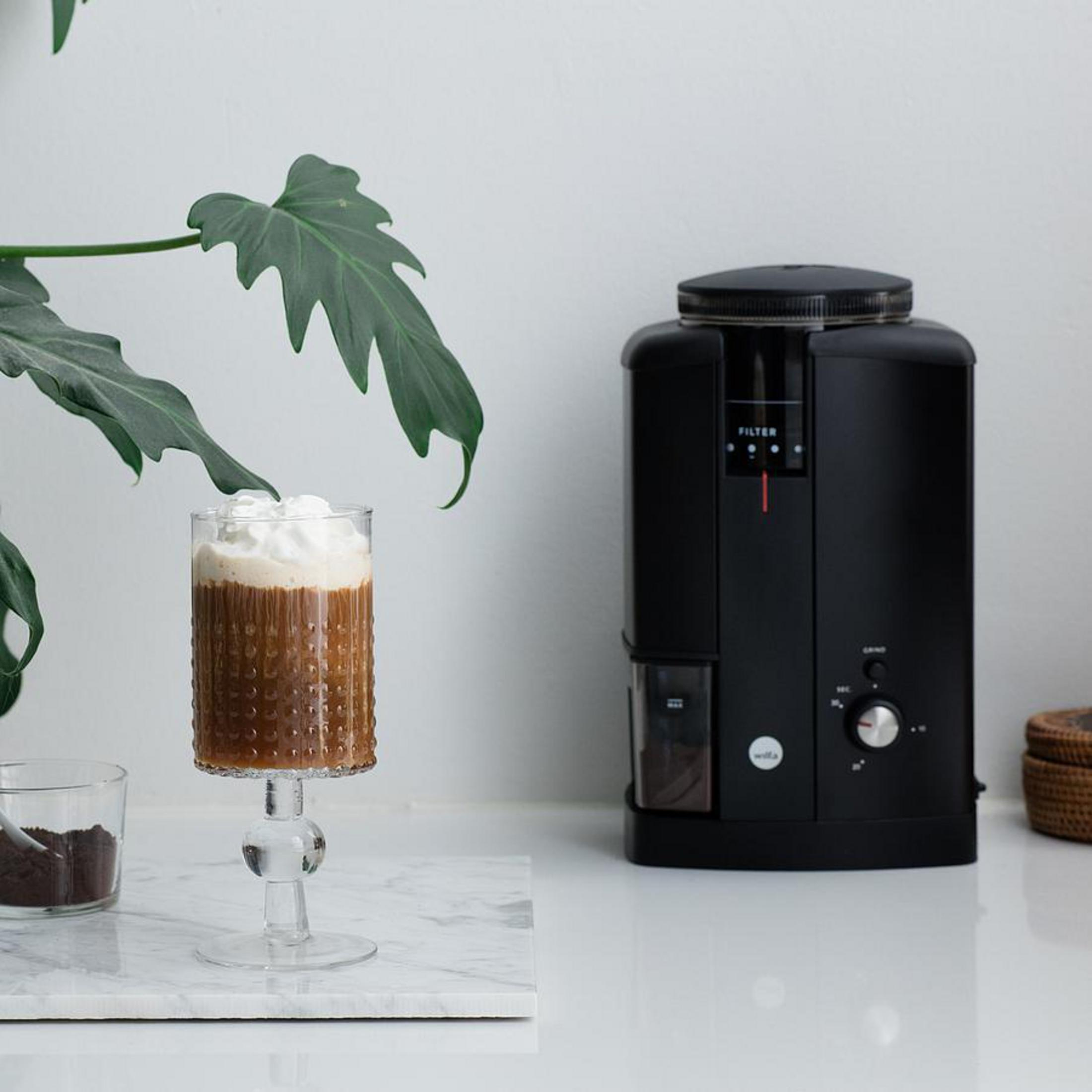 Un bicchiere di caffè con panna si trova accanto a un macinacaffè e una pianta.
