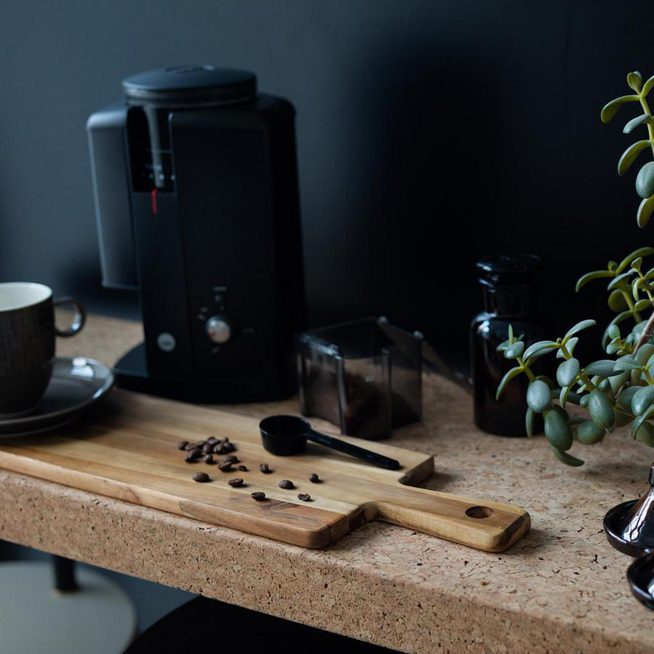 Macinacaffè nero, tazza, chicchi di caffè su tagliere, bottiglia di vetro e pianta.