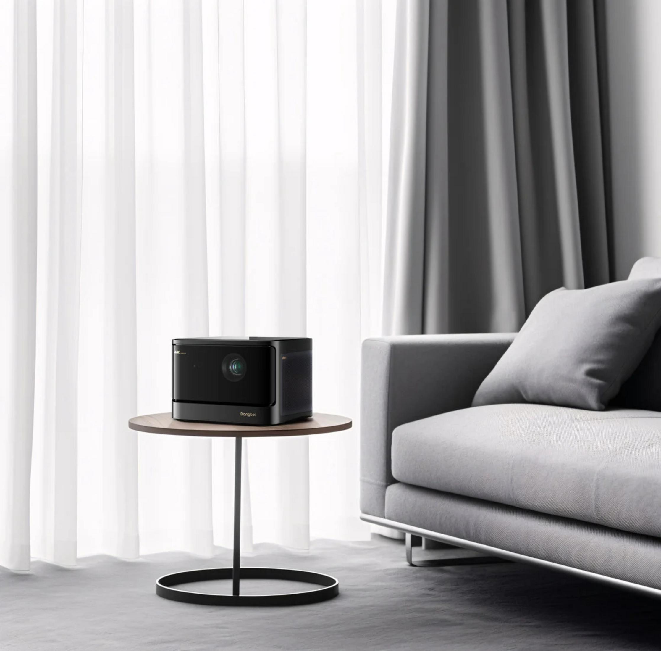Un projecteur noir est sur une table ronde à côté d'un canapé gris avec des rideaux blancs derrière.