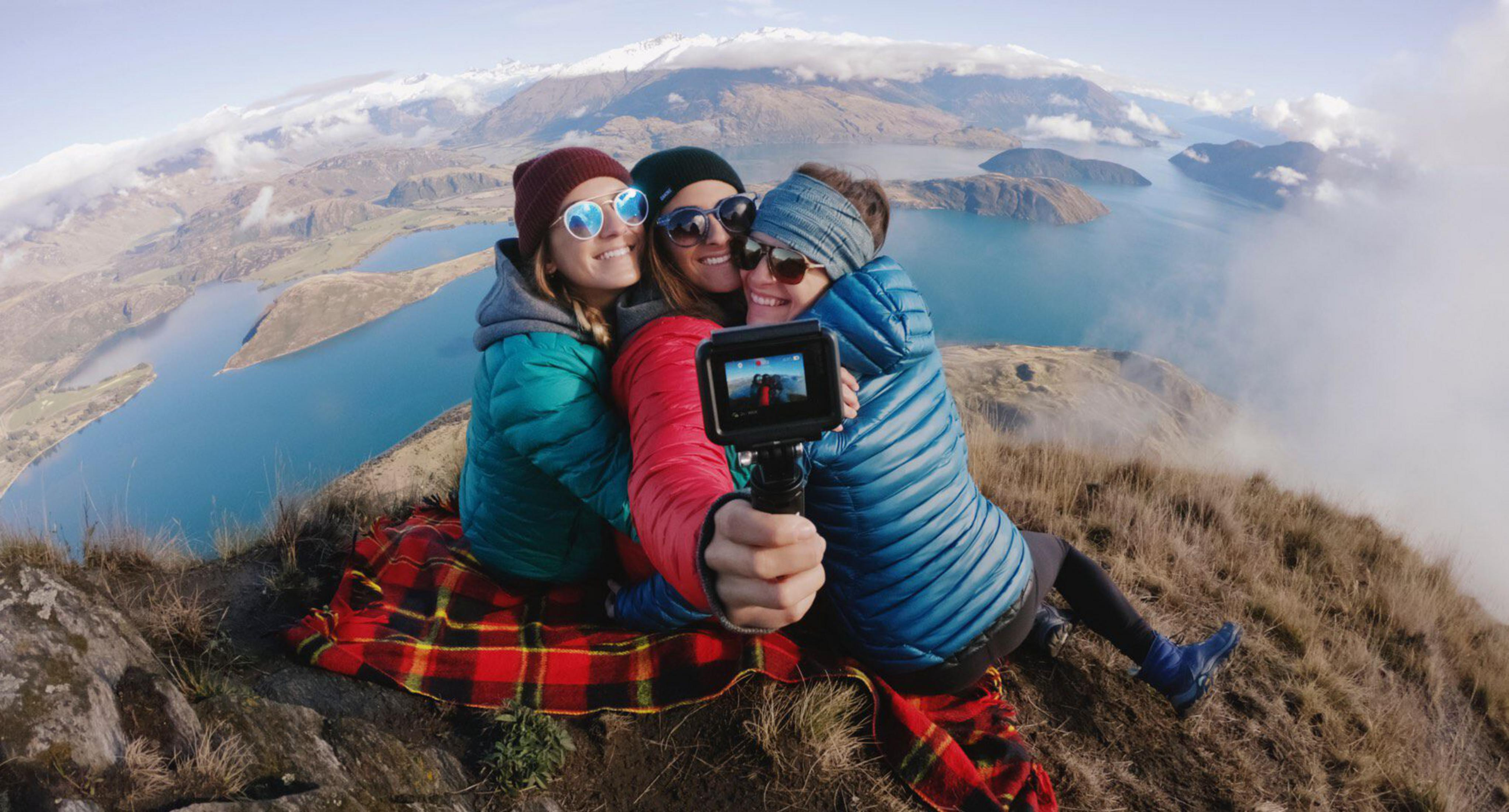 Tre donne si scattano un selfie su una montagna con vista su un lago e montagne innevate.