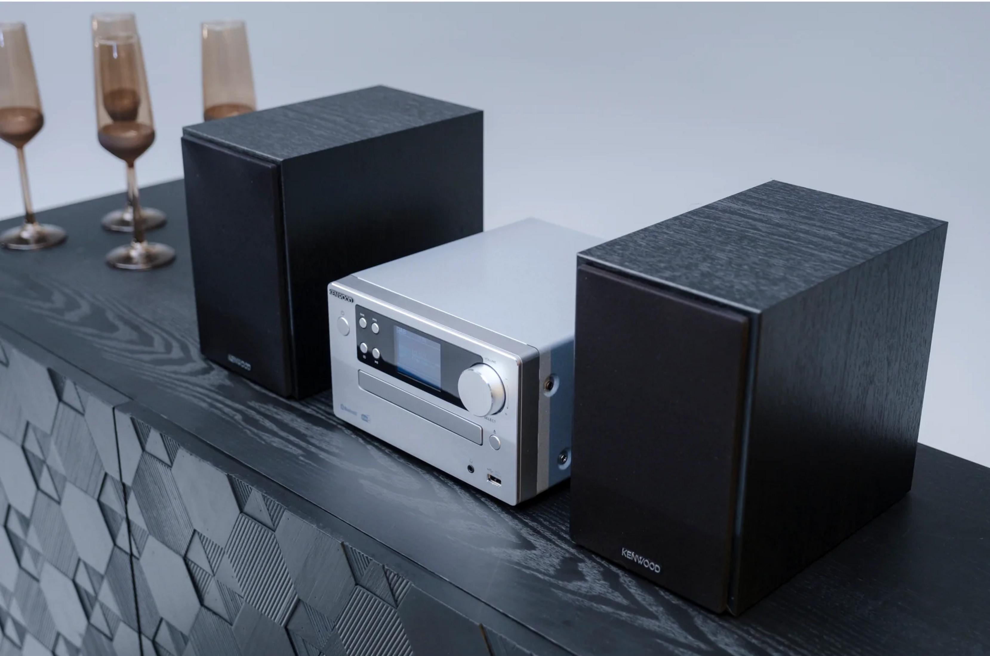 Ricevitore CD Kenwood argento tra due altoparlanti neri su un tavolo scuro.