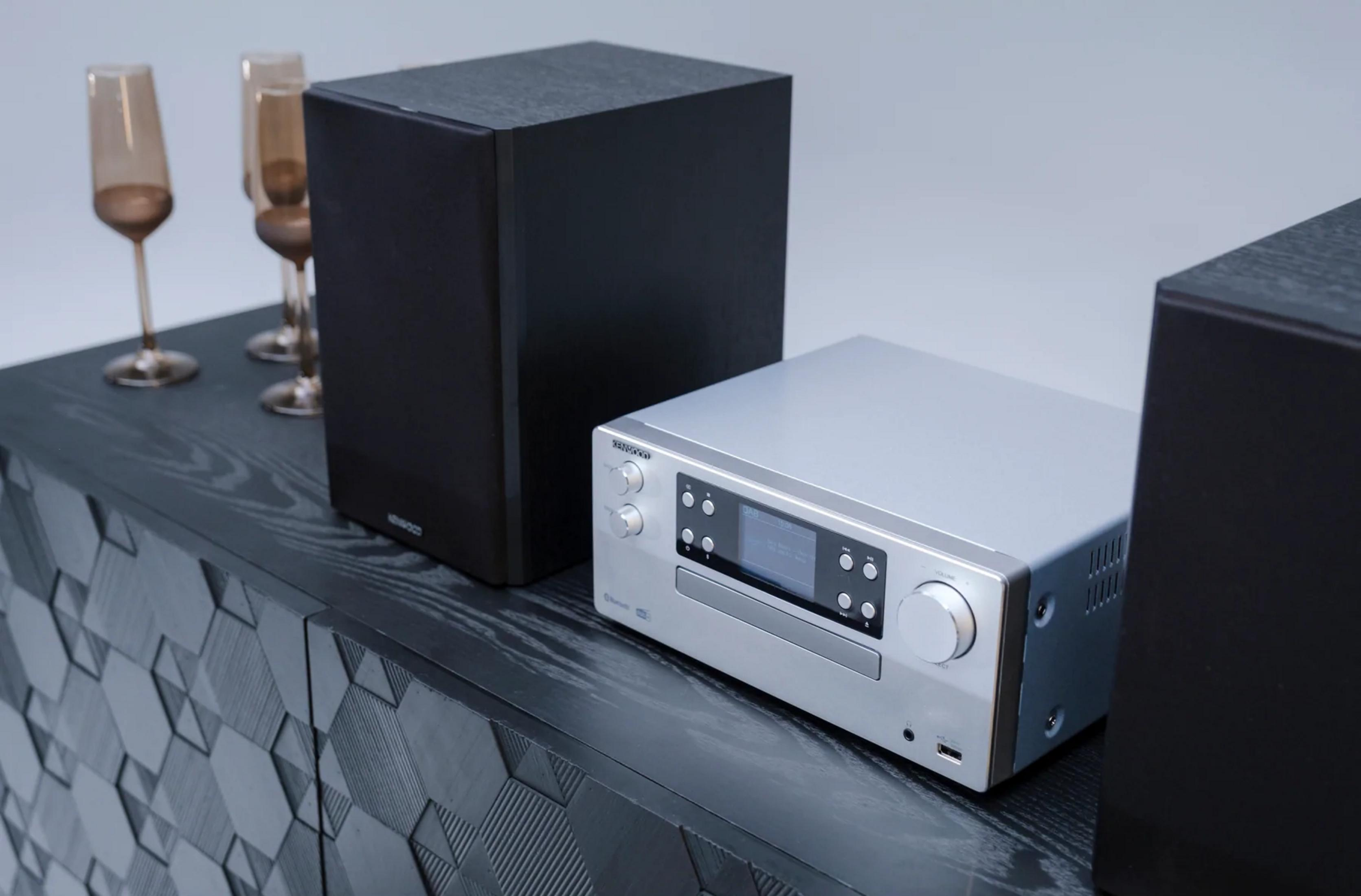 Silberner Kenwood Receiver mit zwei schwarzen Lautsprechern auf einem dunklen Schrank.