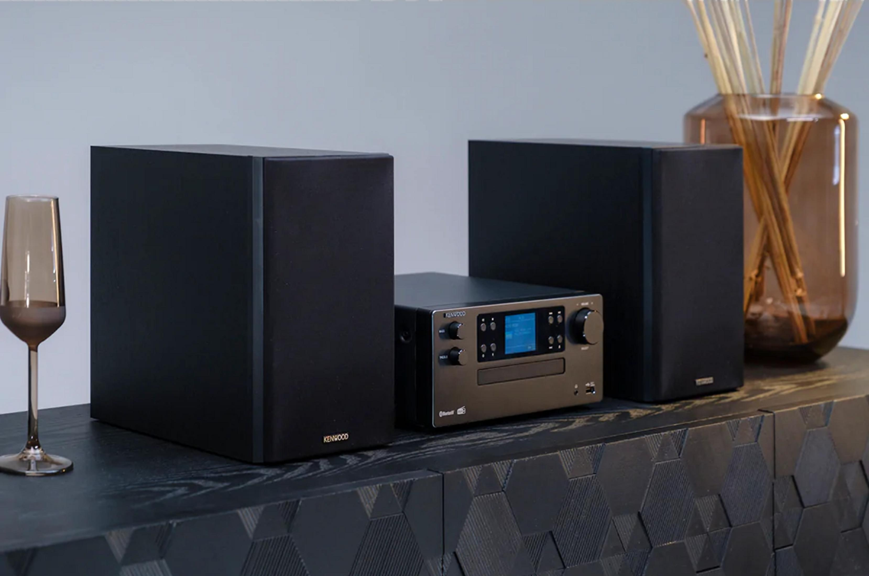 Ein Kenwood Hi-Fi-System mit zwei Lautsprechern, Receiver und Glas auf einem dunklen Schrank.
