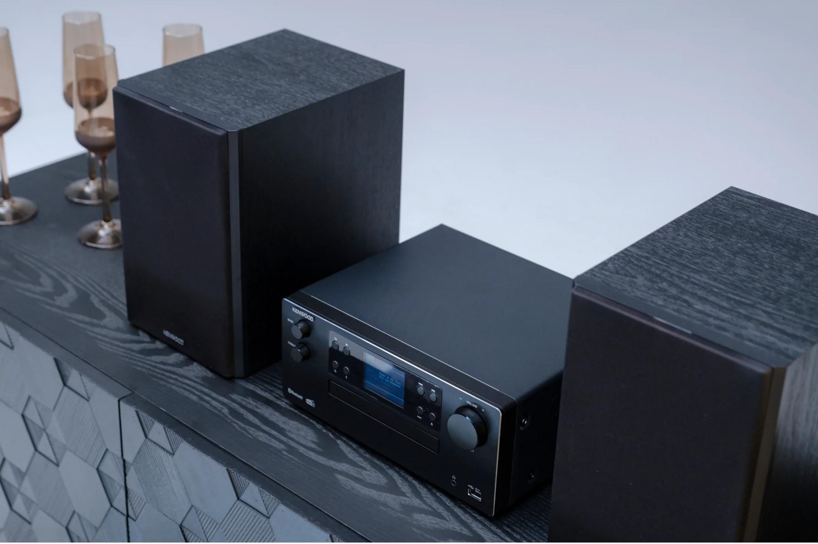 Schwarzes Stereo-System mit zwei Lautsprechern und einer Haupteinheit auf einem dunklen Schrank.