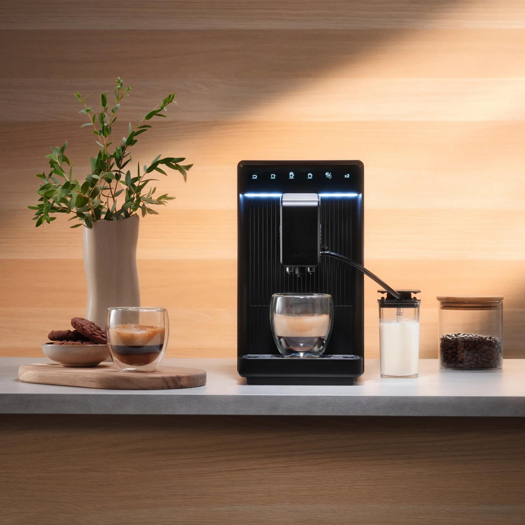 Cafetera prepara espresso en vaso, con leche, granos de café y galletas cerca.