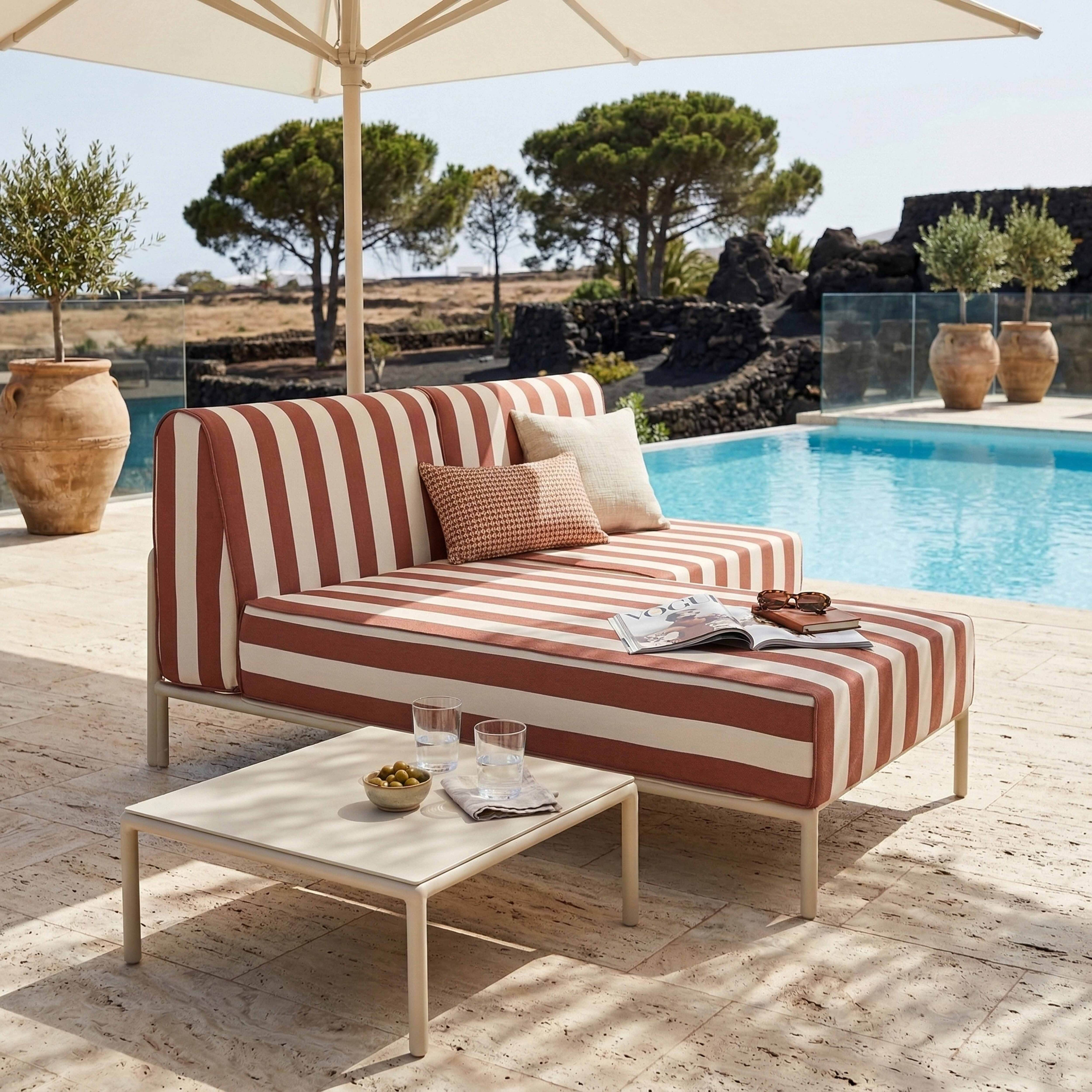 Gestreifte Couch mit Kissen und Beistelltisch am Pool. Olivenbaum im Topf.