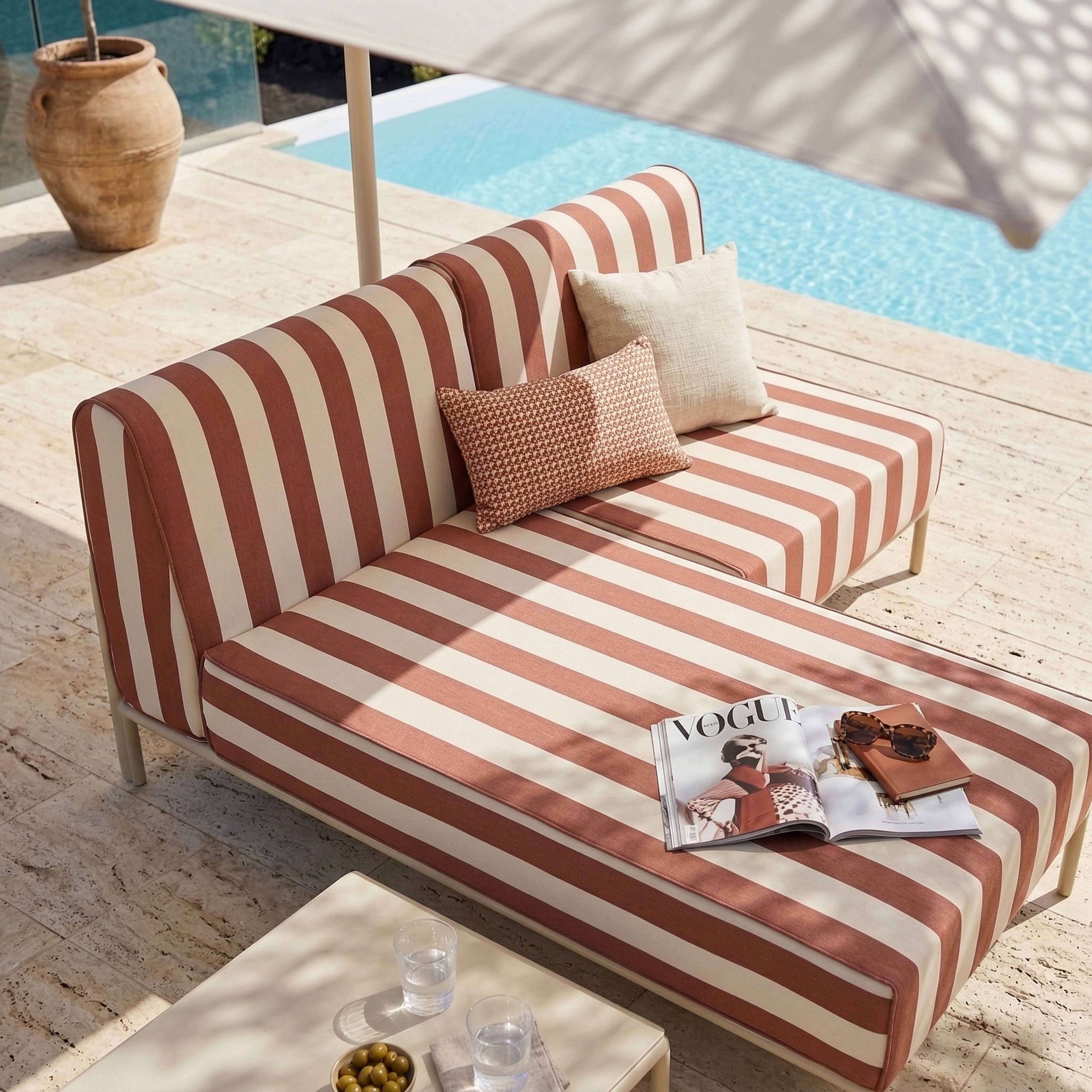 Ein gestreiftes Outdoor-Sofa am Pool mit einer Zeitschrift und Sonnenbrille.