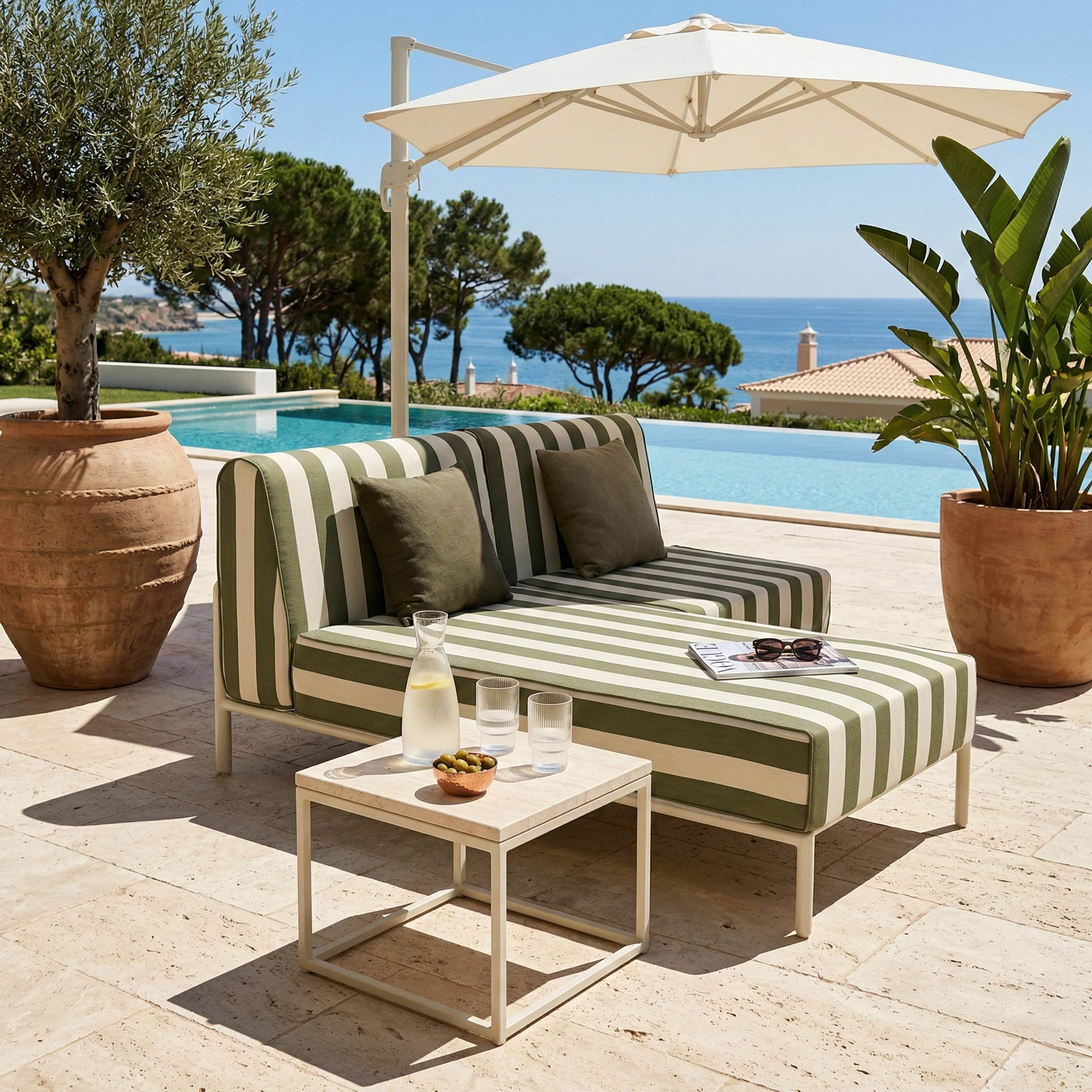 Gestreifte Couch am Pool mit Oliven, Getränken und Sonnenbrille. Bäume und Meer im Hintergrund.