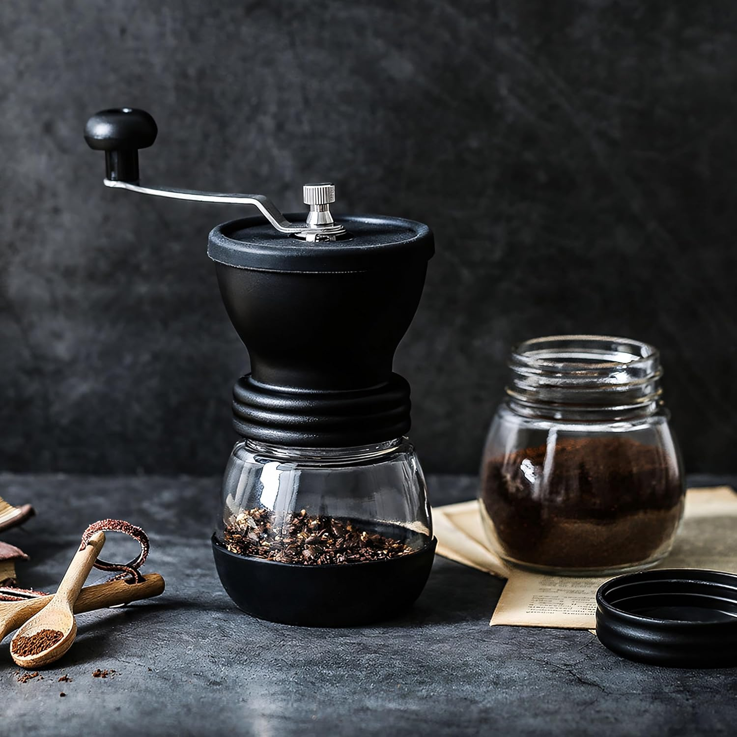 Eine schwarze Kaffeemühle mit Glasbehälter steht neben einem Glas mit Kaffeepulver.