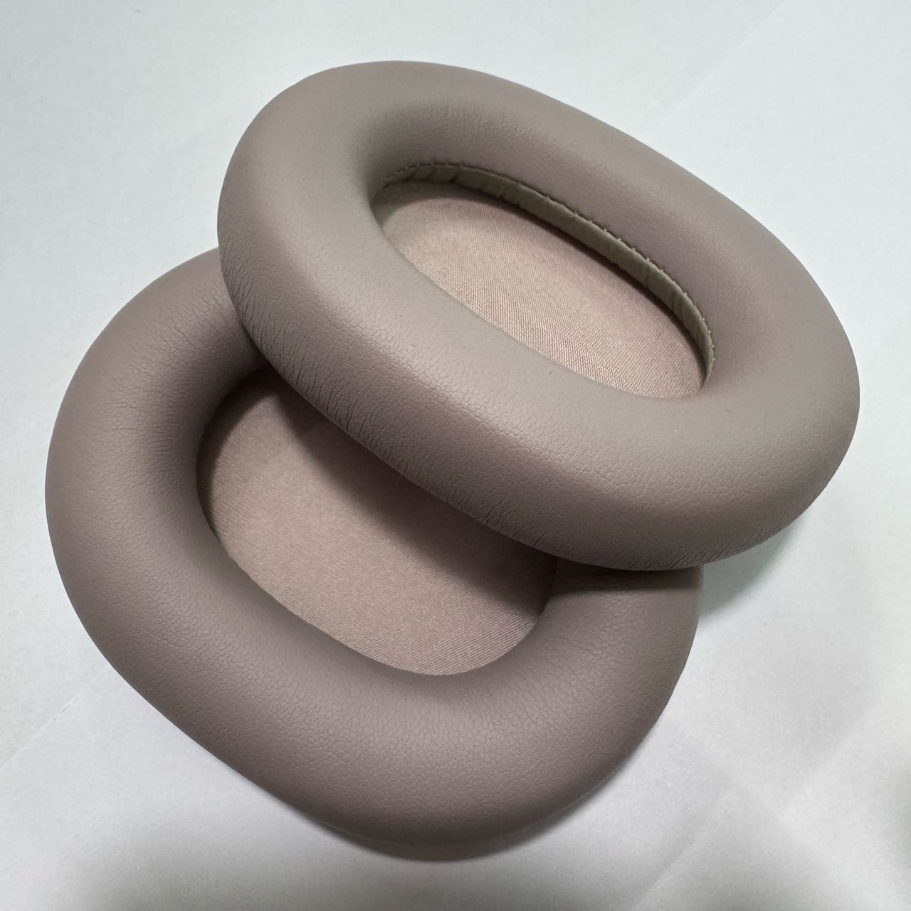 Due cuscinetti auricolari ovali color tortora sono sovrapposti su sfondo bianco.