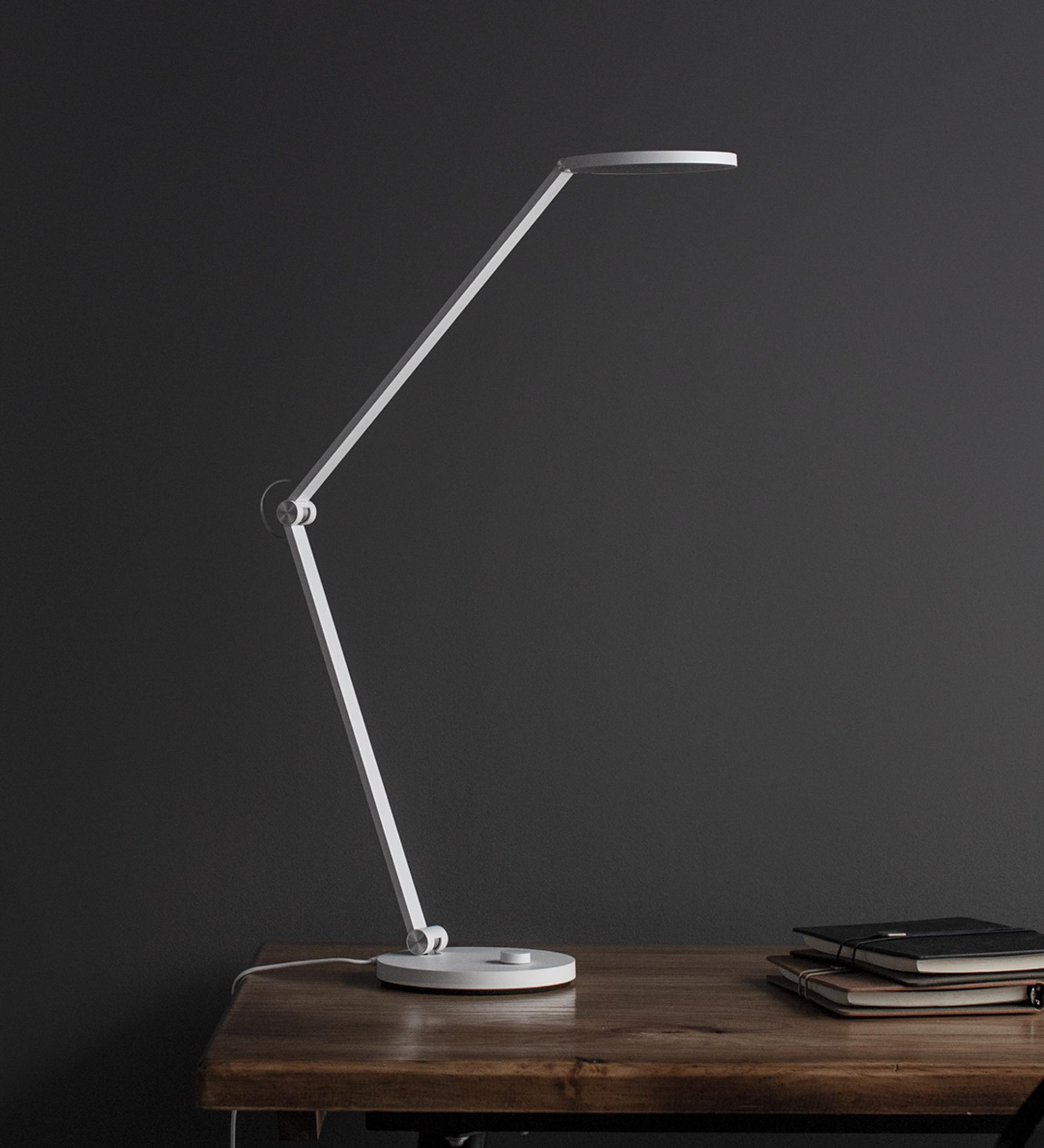 Biała lampa biurkowa z regulowanym ramieniem stoi na drewnianym stole obok notatników.