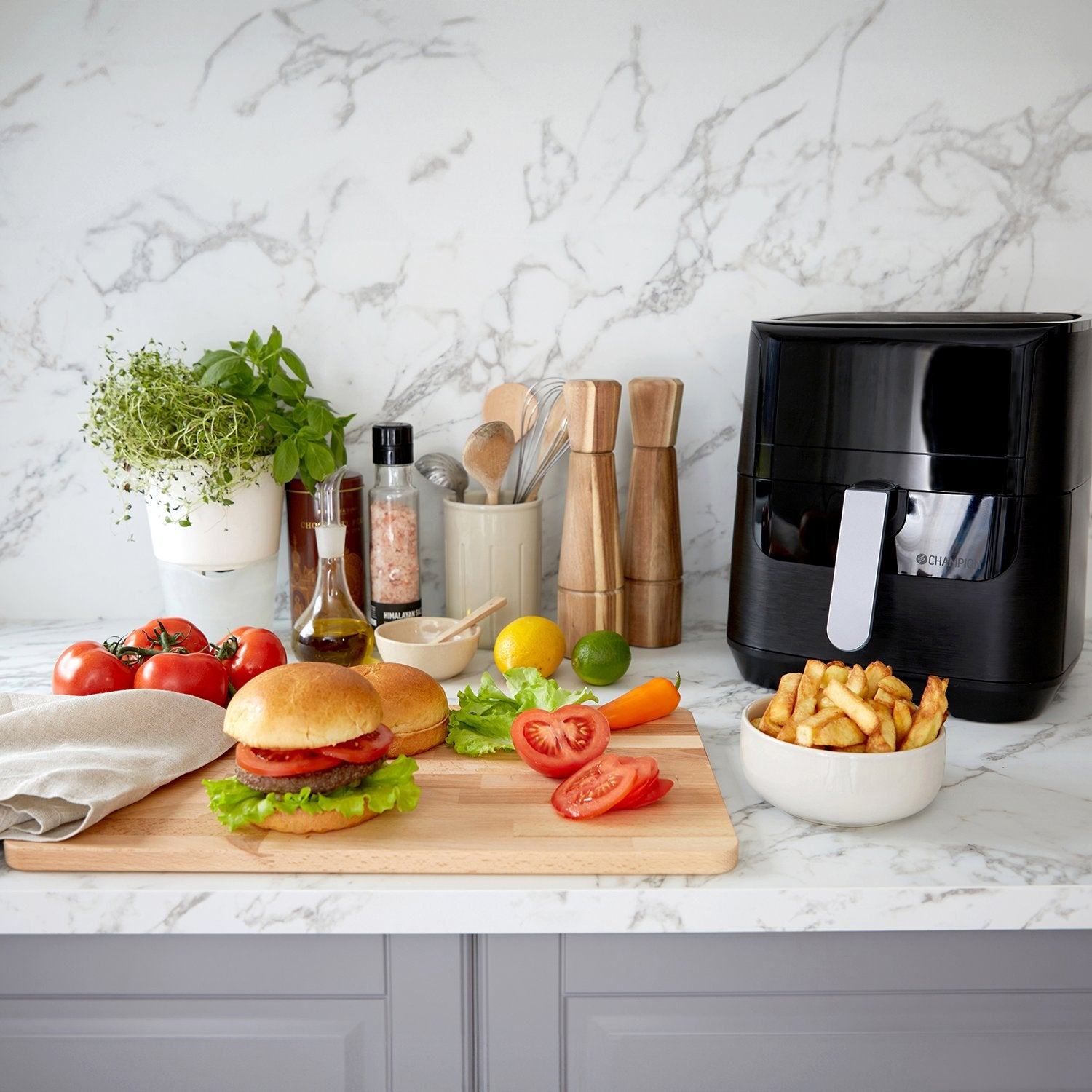 A burger on a cutting board with tomatoes, lettuce, and fries next to an air fryer.