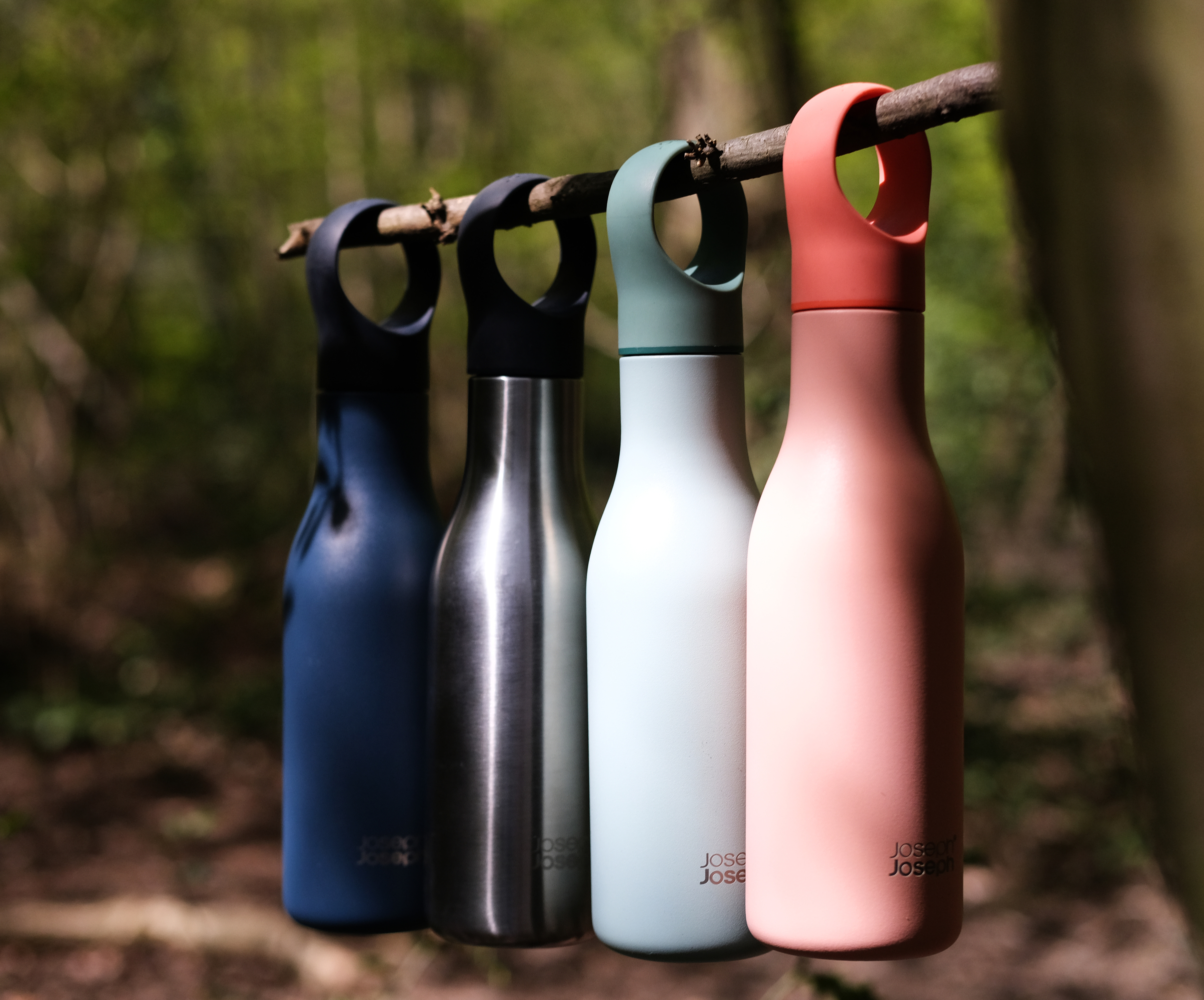 Vier Wasserflaschen in Blau, Silber, Hellgrün und Rosa hängen an einem Ast.