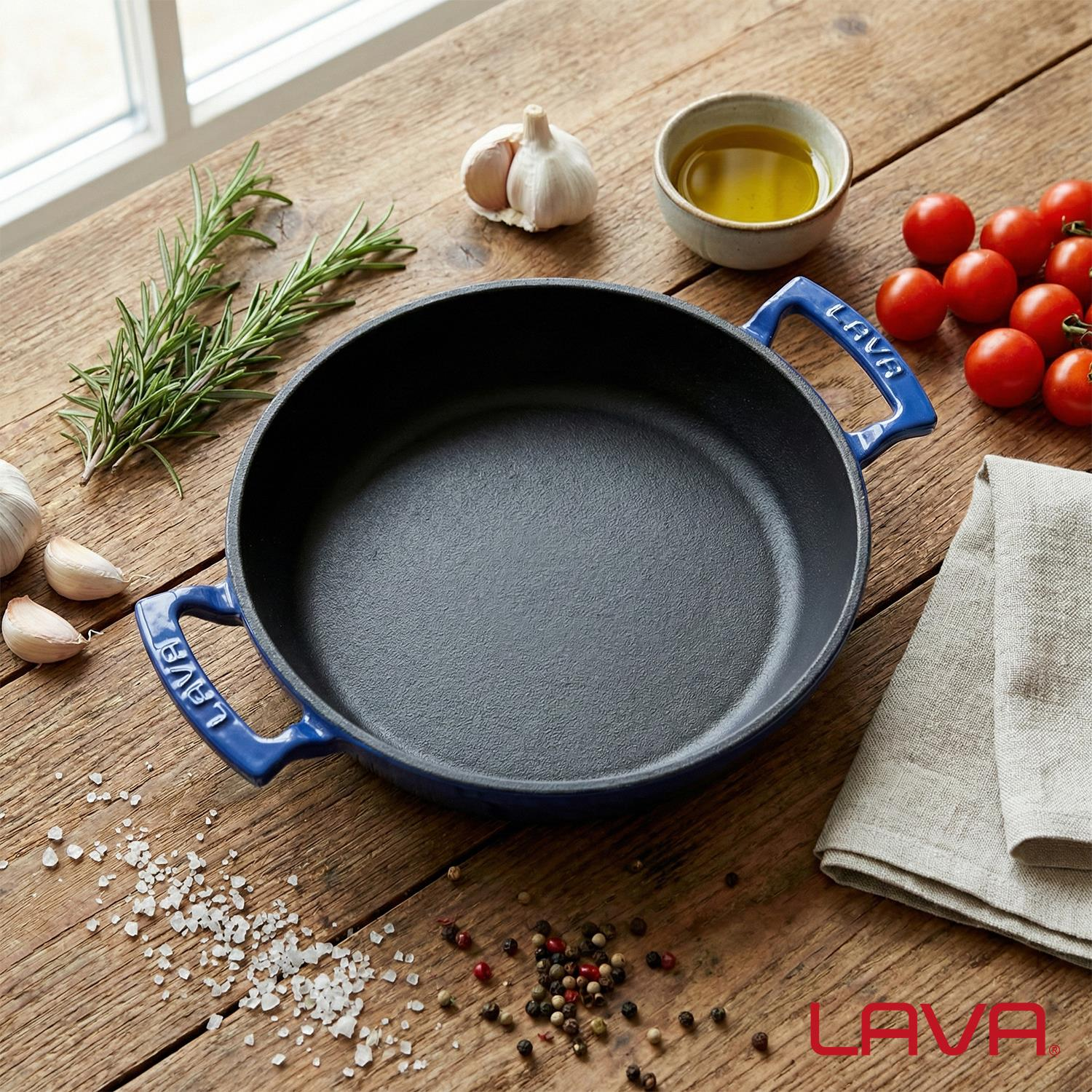 Mavi kulplu döküm tencere, biberiye, sarımsak, zeytinyağı ve domateslerle çevrili.