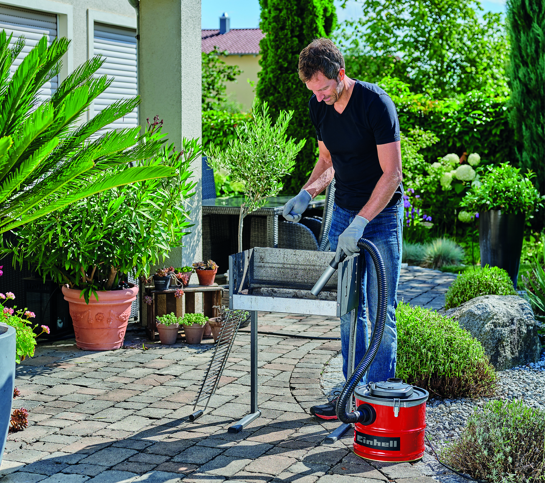 Ein Mann reinigt einen Grill mit einem roten Aschesauger in einem Garten.