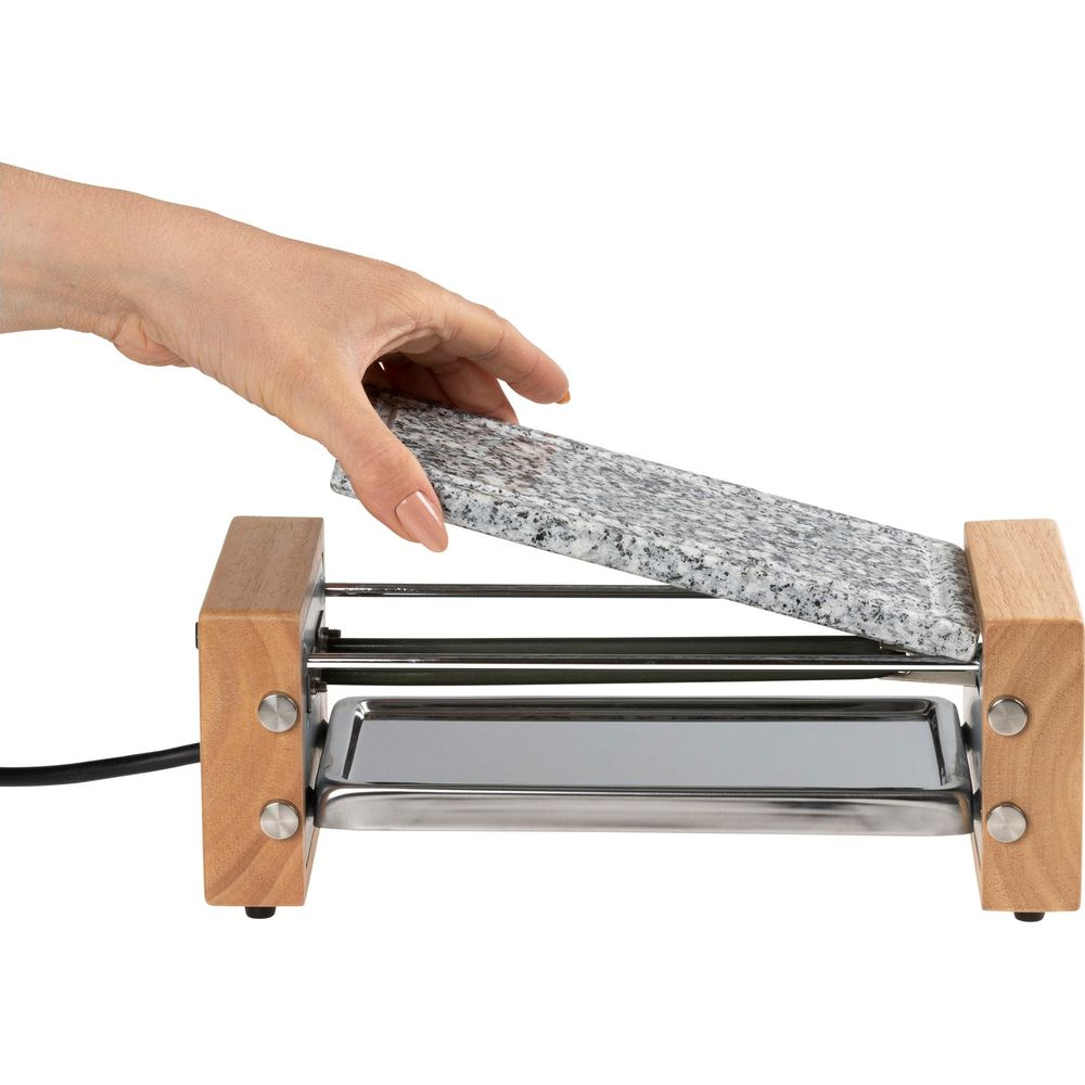 Eine Hand legt eine Granitplatte auf ein Raclettegerät aus Holz und Metall.