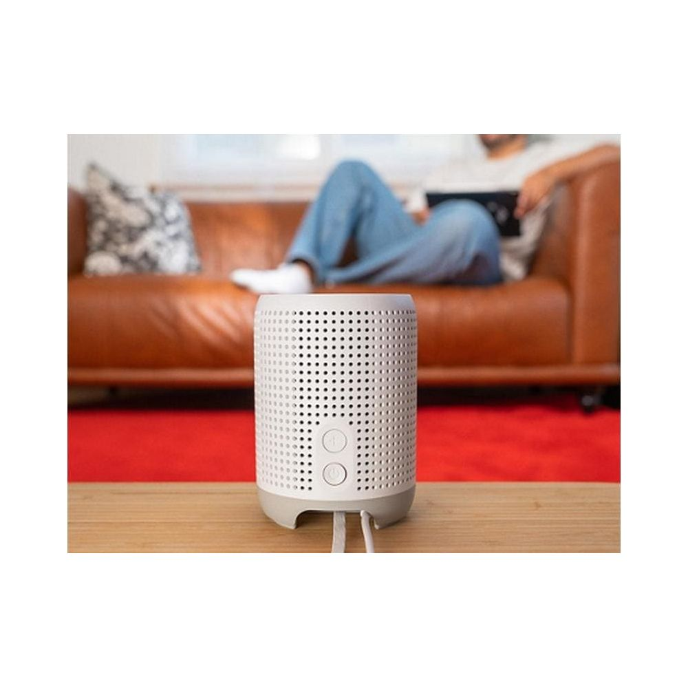 Une enceinte blanche sur une table en bois. Une personne est allongée sur un canapé.