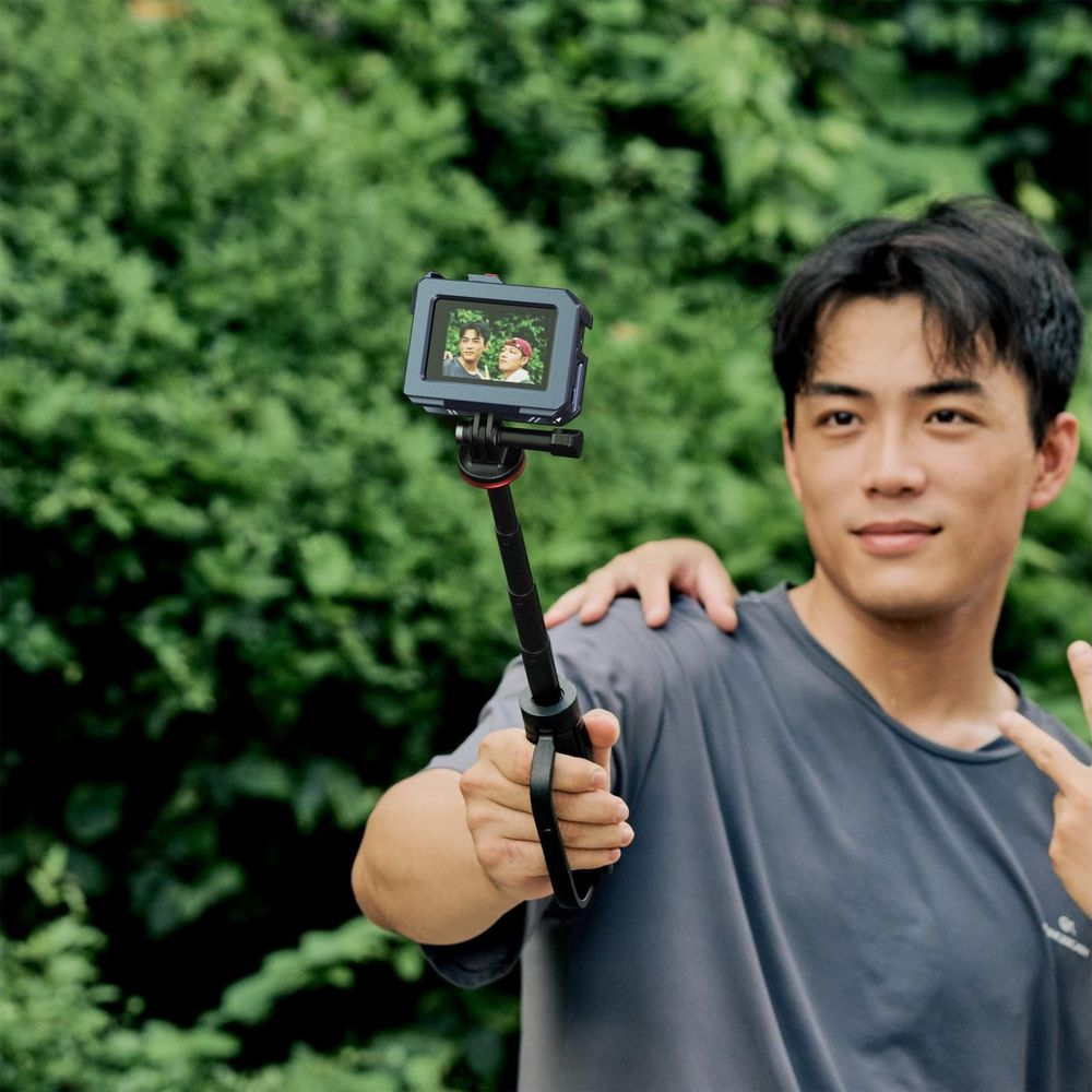 Un homme prend un selfie avec une caméra sur une perche à selfie devant un feuillage vert.