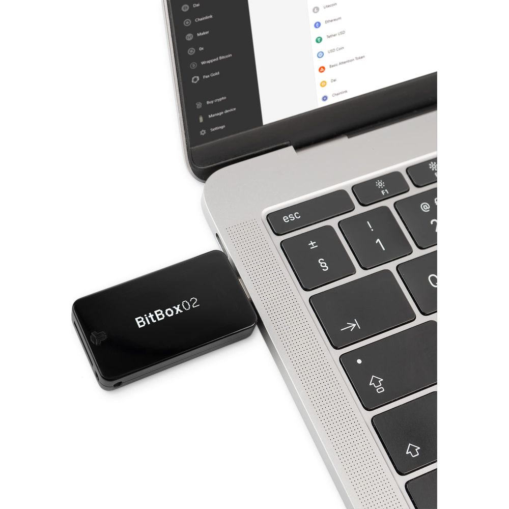 Un dispositivo BitBox02 nero è collegato a un laptop. Il laptop mostra un elenco di criptovalute.